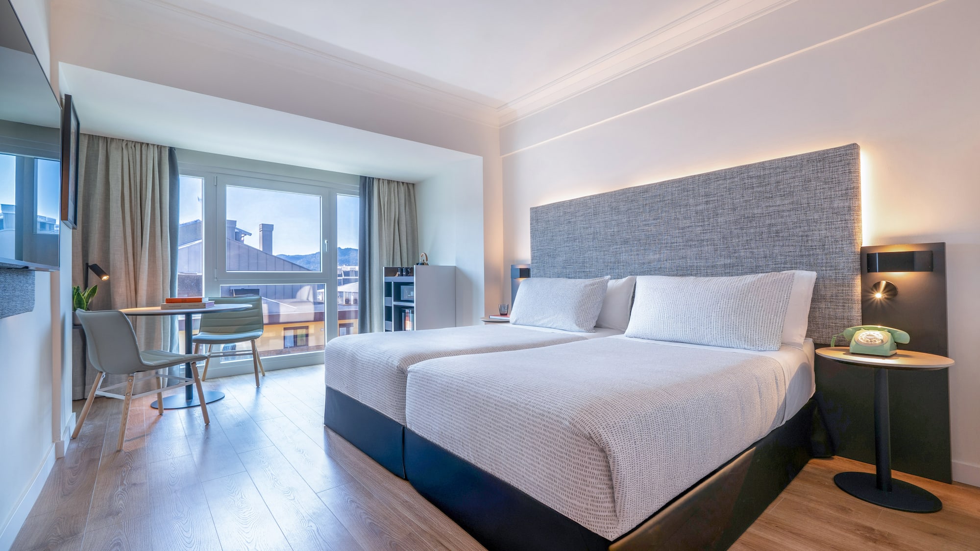 Moderna habitación de hotel con dos camas, grandes ventanas y vista a la ciudad.