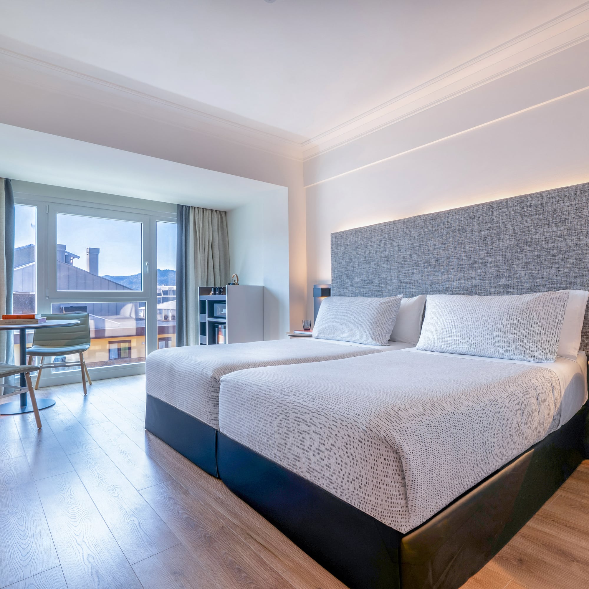 Moderna habitación de hotel con dos camas, grandes ventanas y vista a la ciudad.