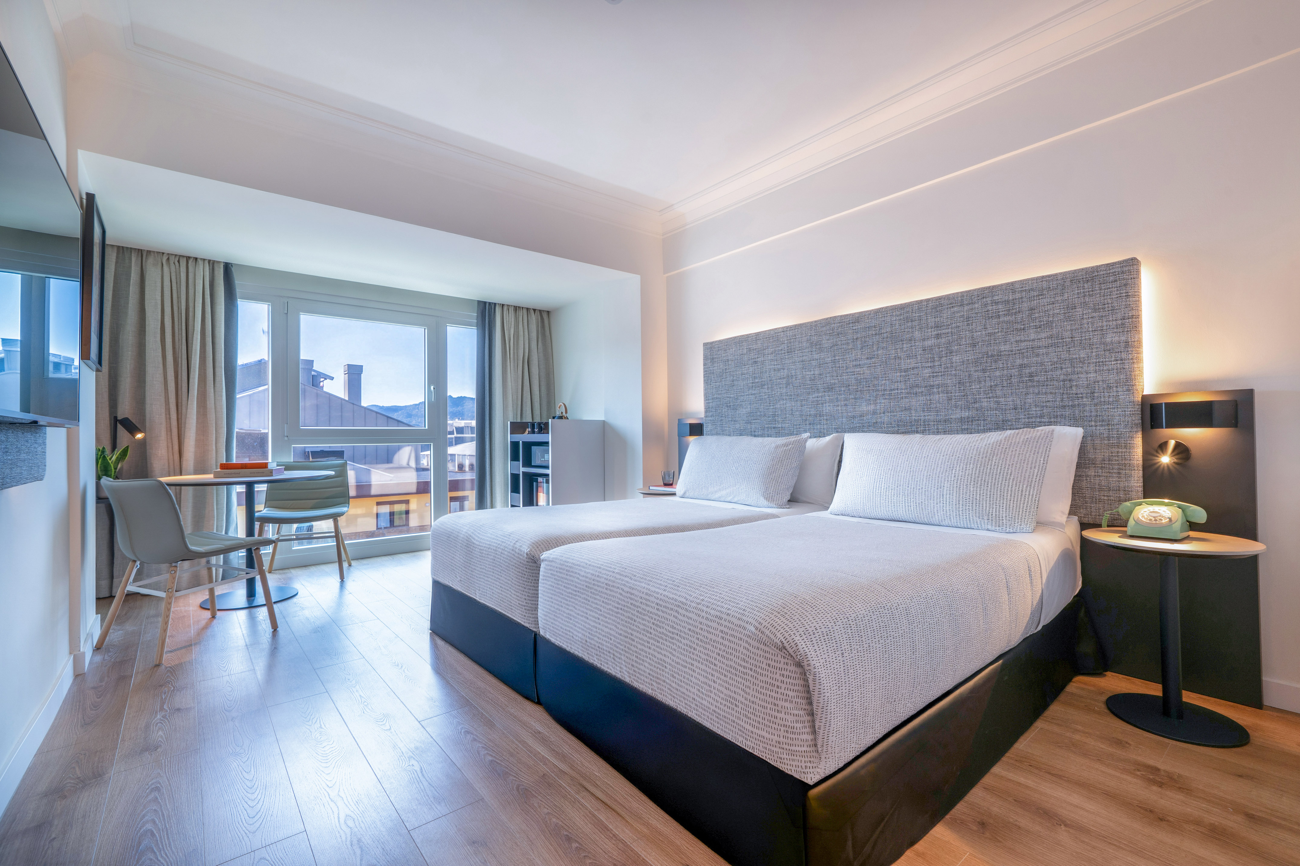 Moderna habitación de hotel con dos camas, grandes ventanas y vista a la ciudad.
