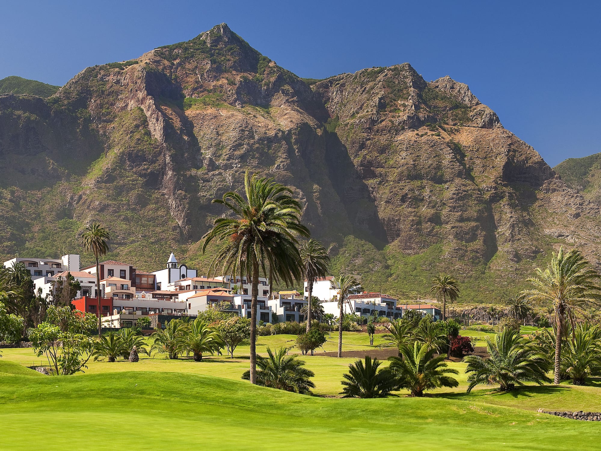 Lush golf course, resort buildings, palm trees, and steep mountains.