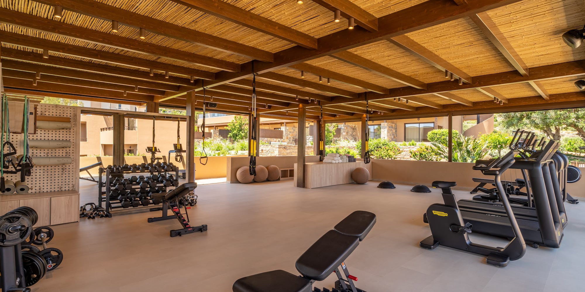 Hotel outdoor gym with bamboo ceiling, weight rack, treadmills, and TRX.