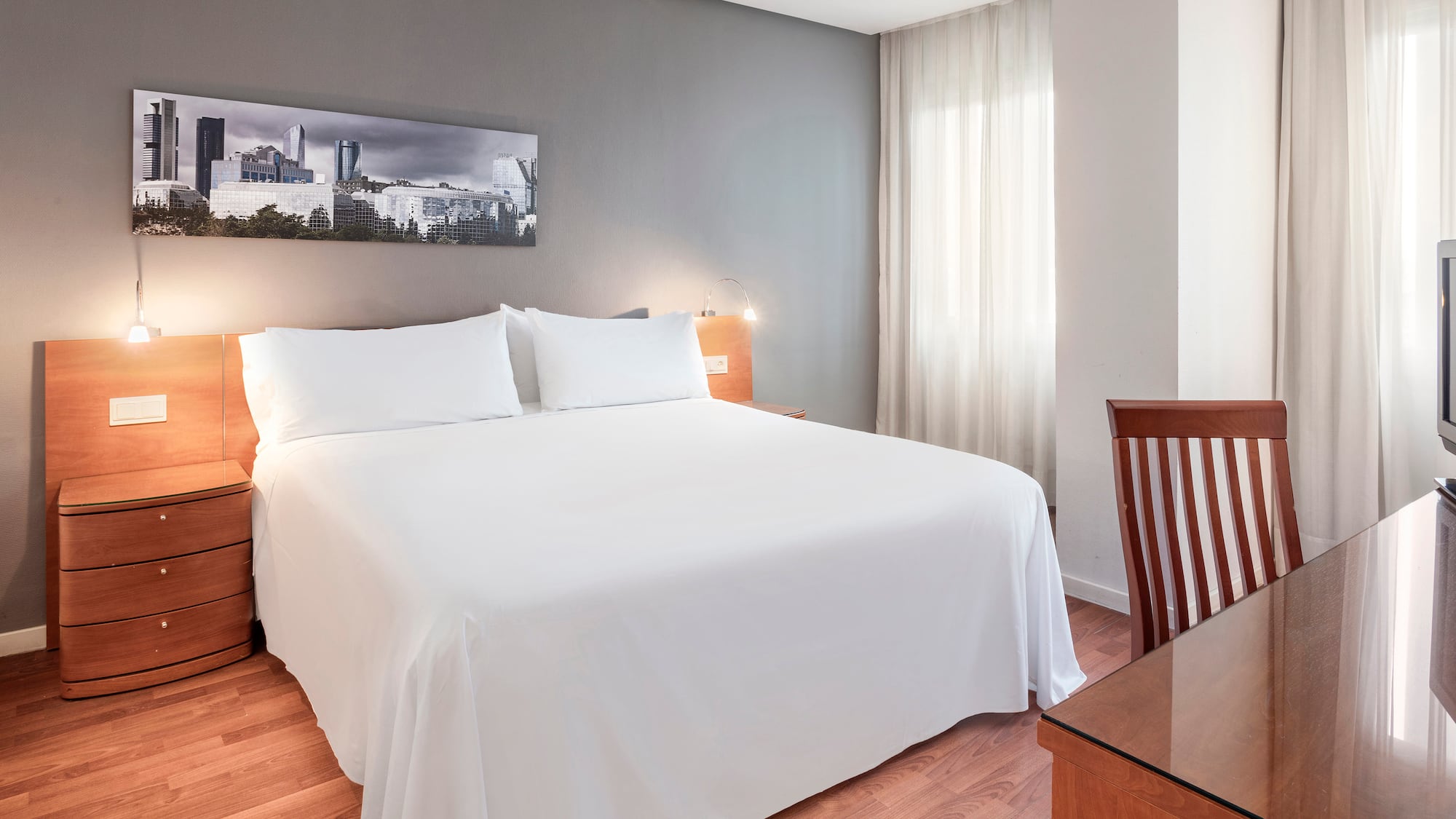 Modern hotel bedroom with white bed, cityscape artwork, and wood flooring.