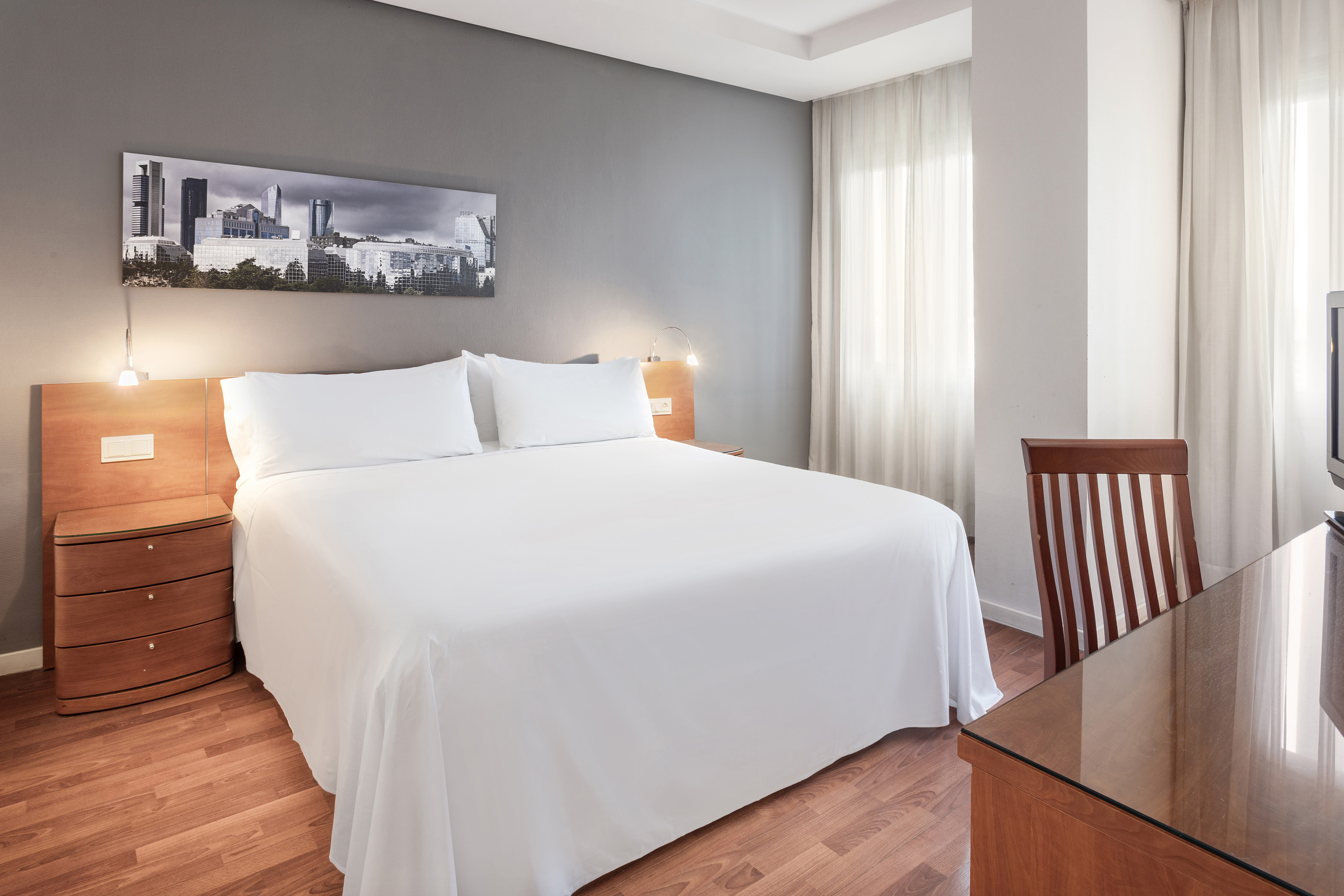 Modern hotel bedroom with white bed, cityscape artwork, and wood flooring.