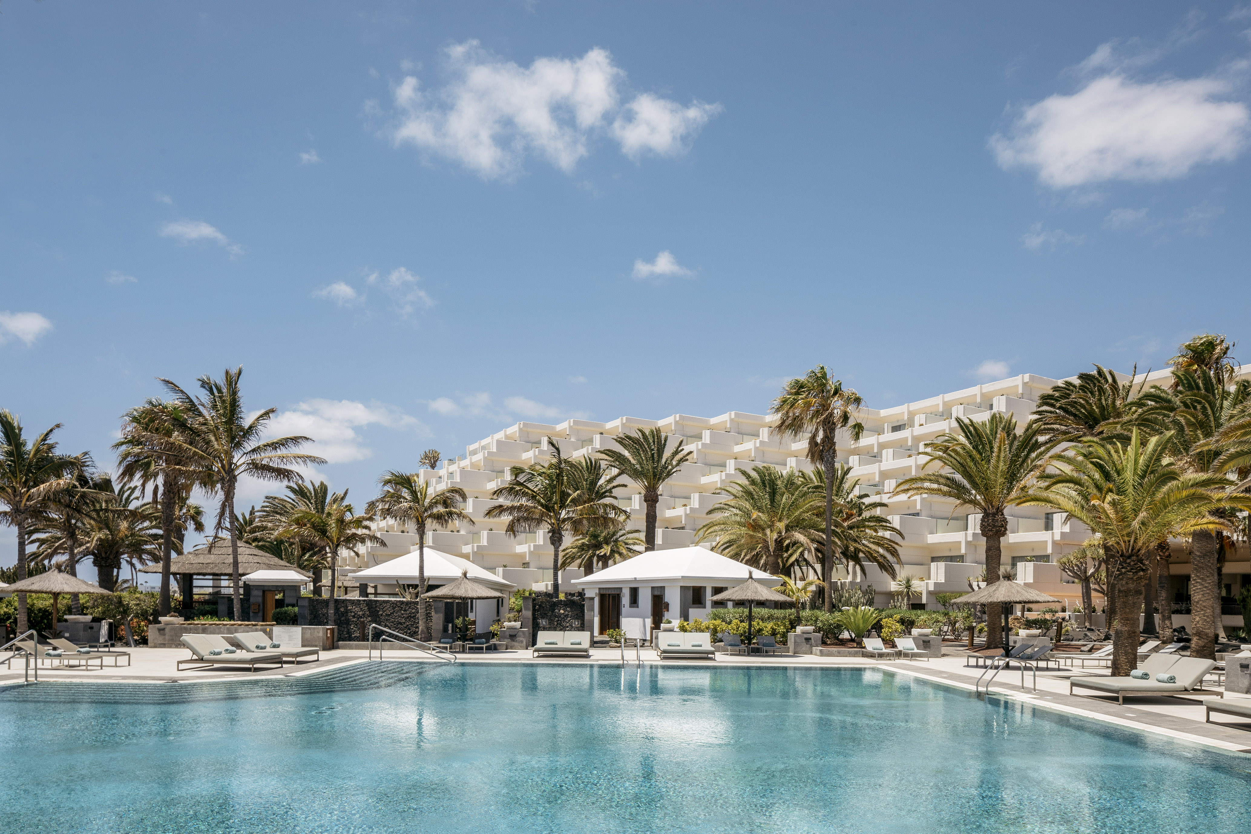 Luxury resort pool with white hotel building and numerous palm trees.