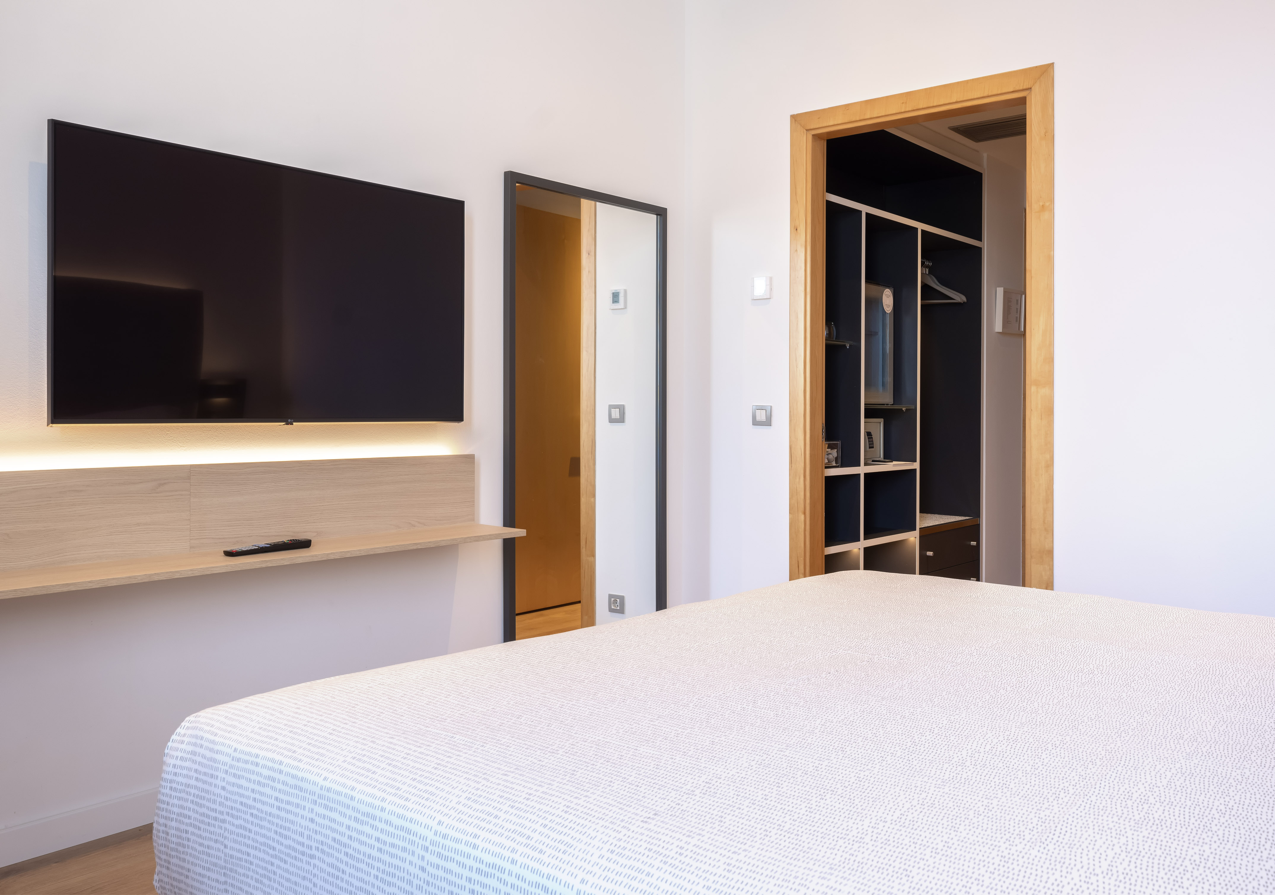 Modern hotel room with wall TV, lit shelf, white bed, mirror, open closet.
