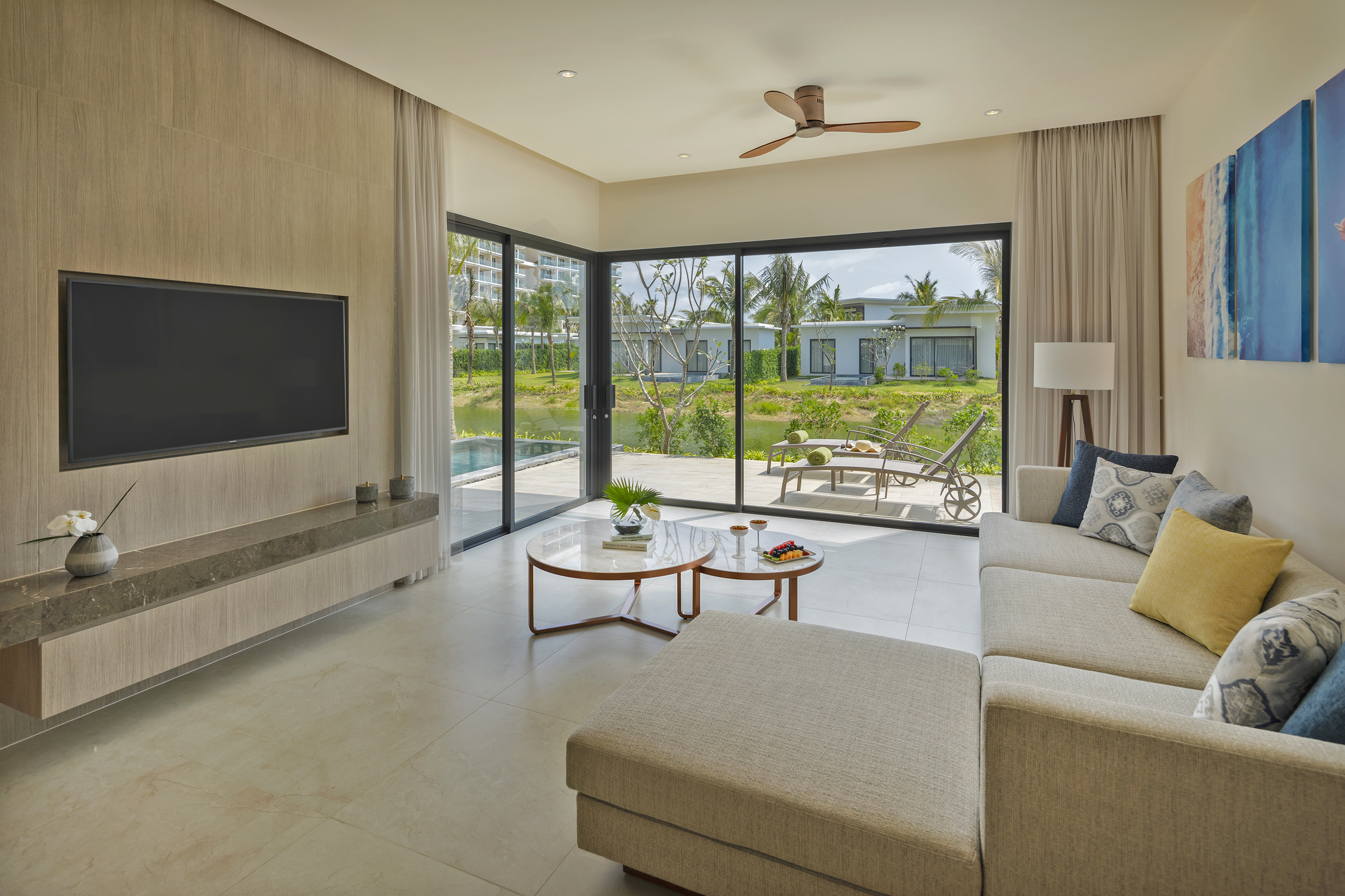 Modern resort living room with sofa, TV, overlooking tropical lake and villas.