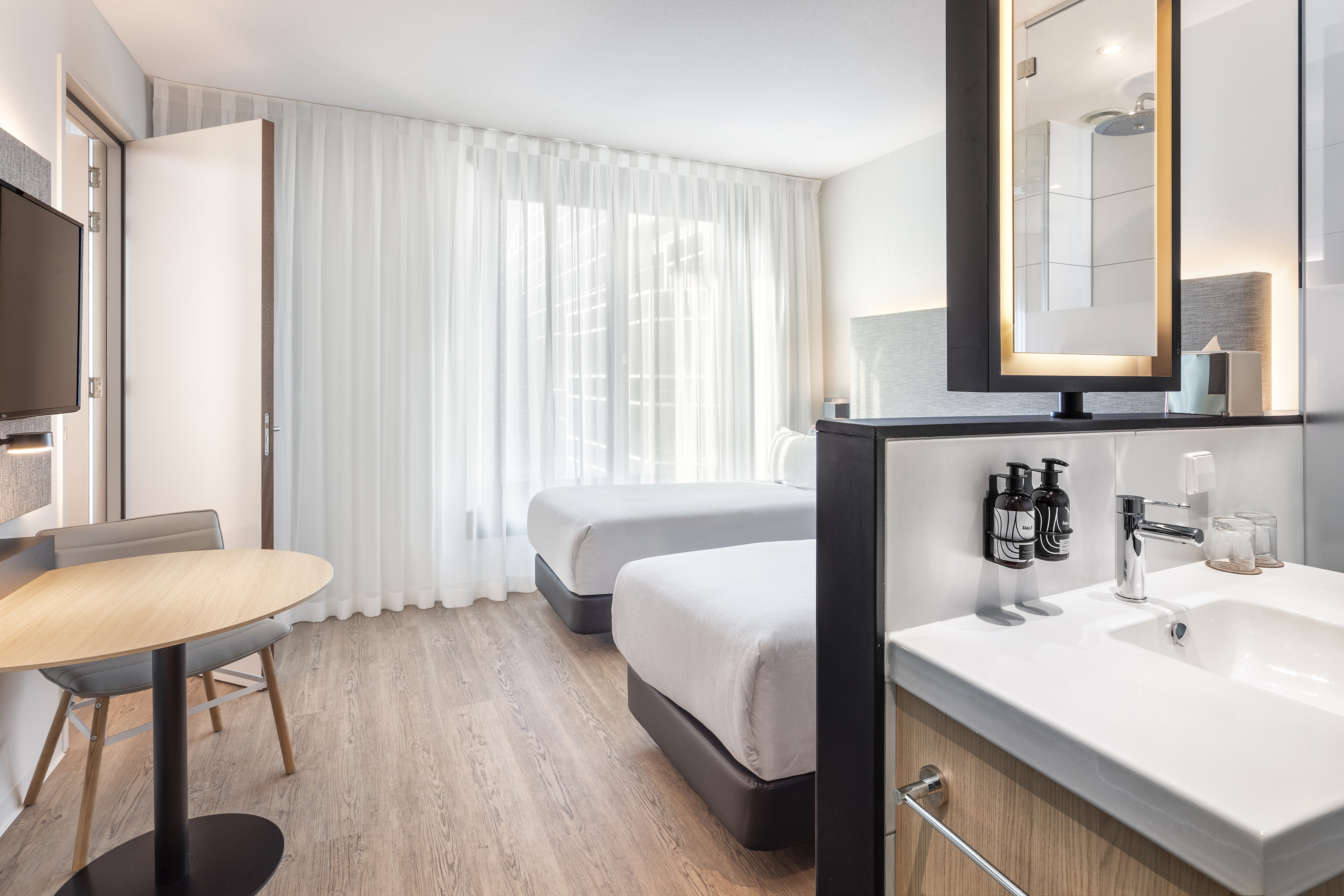 Modern hotel twin room with integrated vanity, wood floor, and sitting area.
