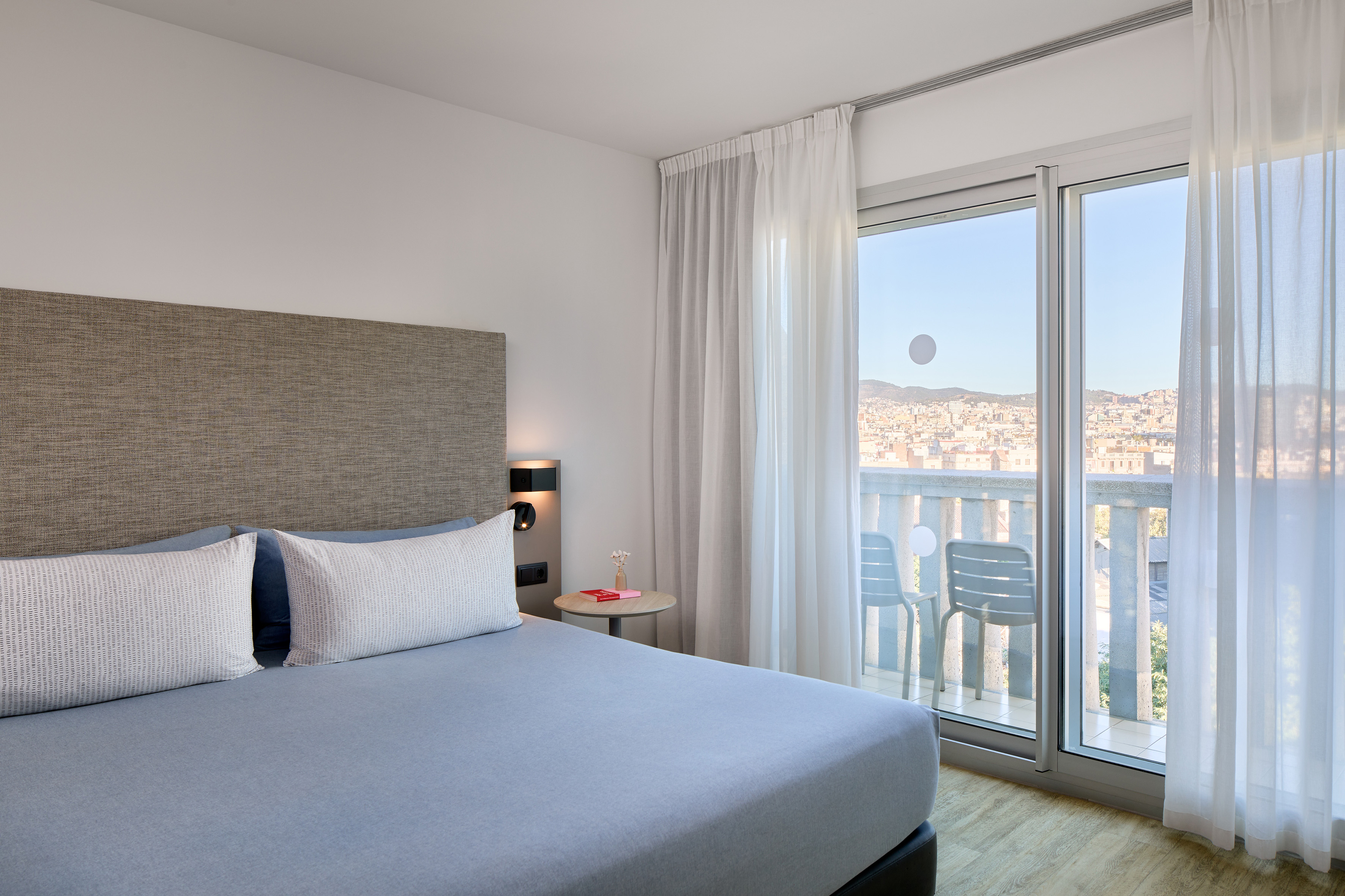 Modern hotel bedroom with bed, nightstand, and balcony overlooking a city.