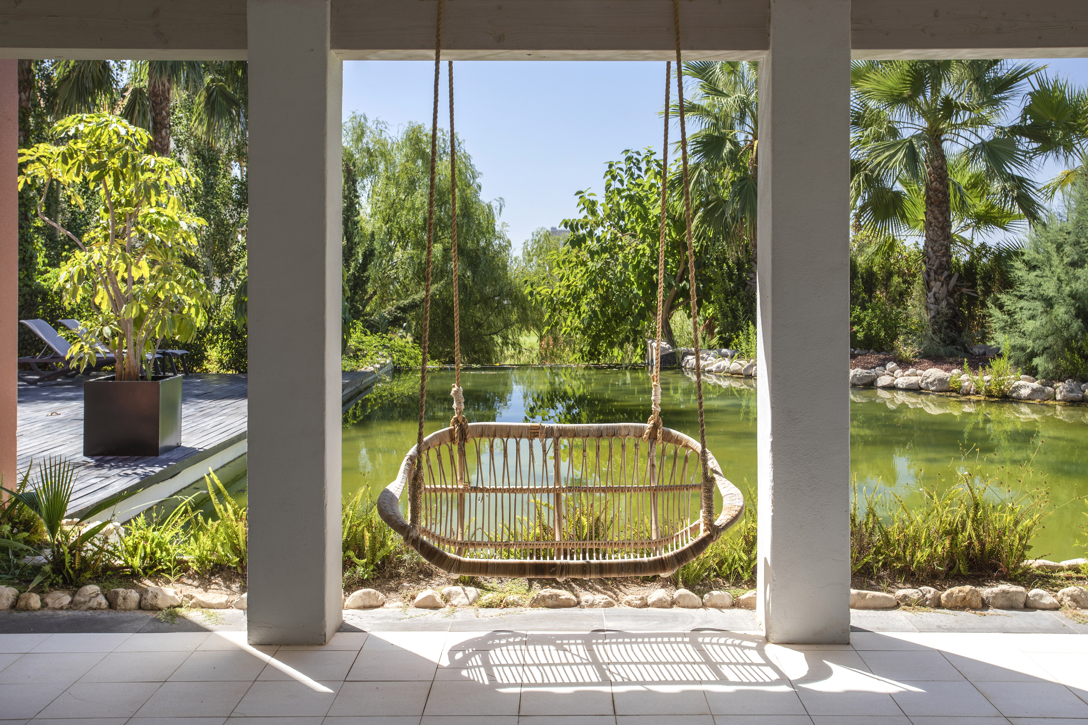 Tropical garden patio with hanging wicker chair overlooking a pond.