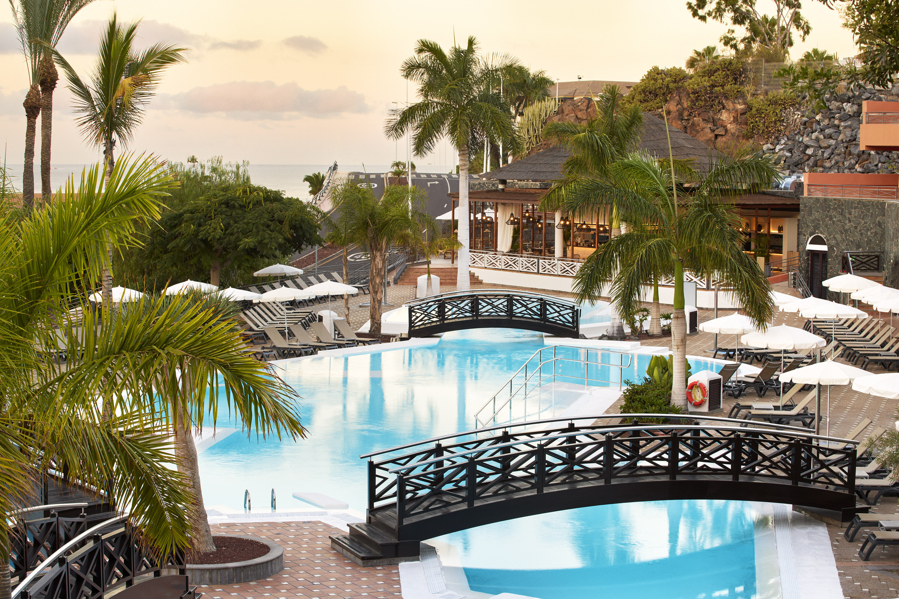 Resort pool with palm trees, sun loungers, and bridges at sunset.