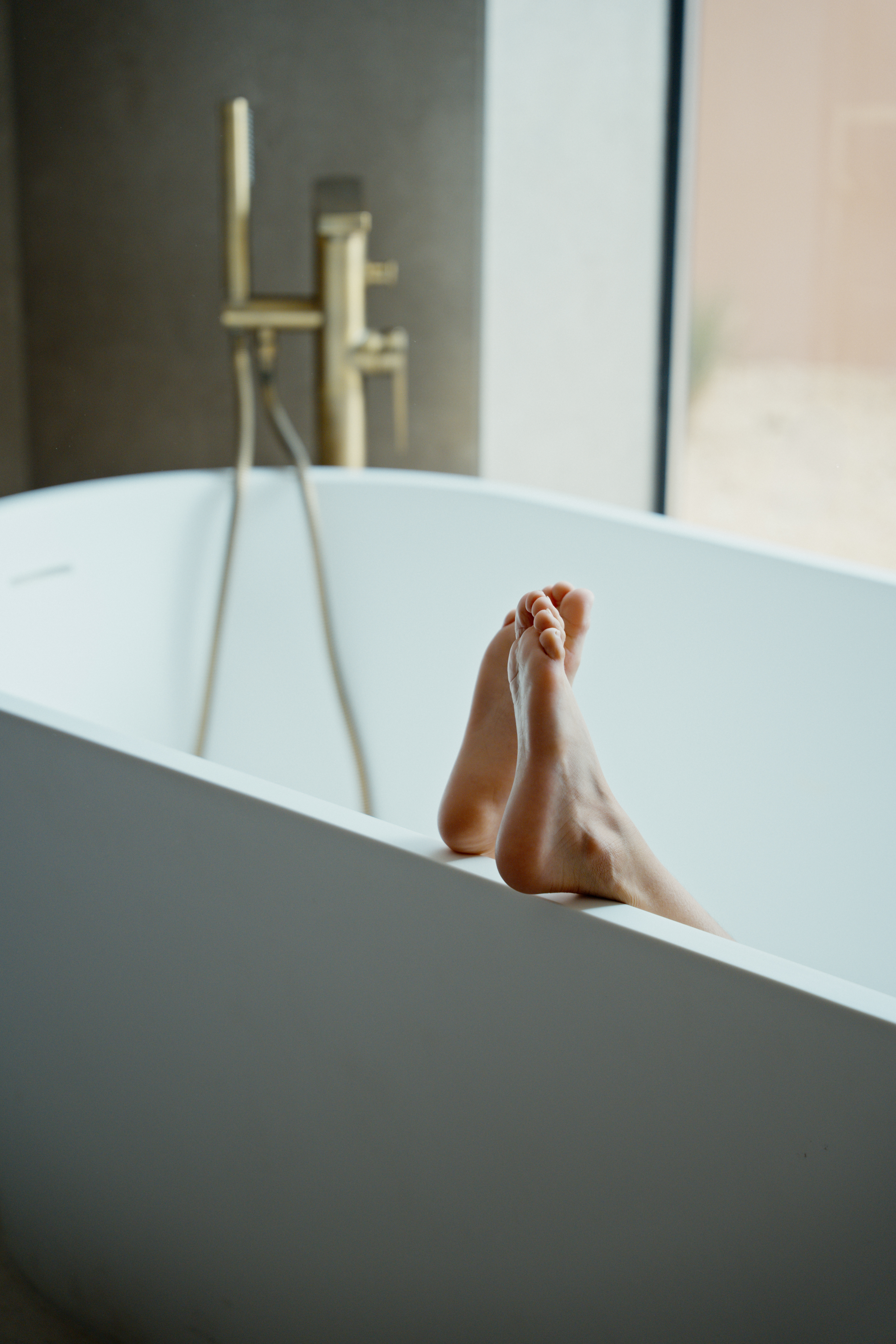 Bare feet on edge of modern white bathtub with golden faucet.