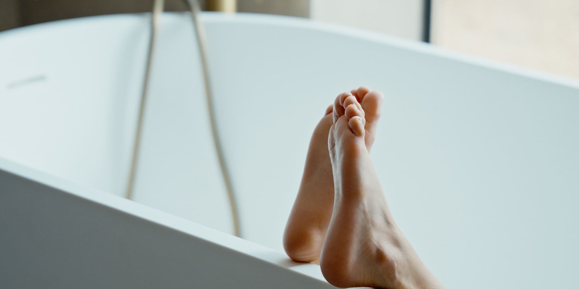 Bare feet on edge of modern white bathtub with golden faucet.