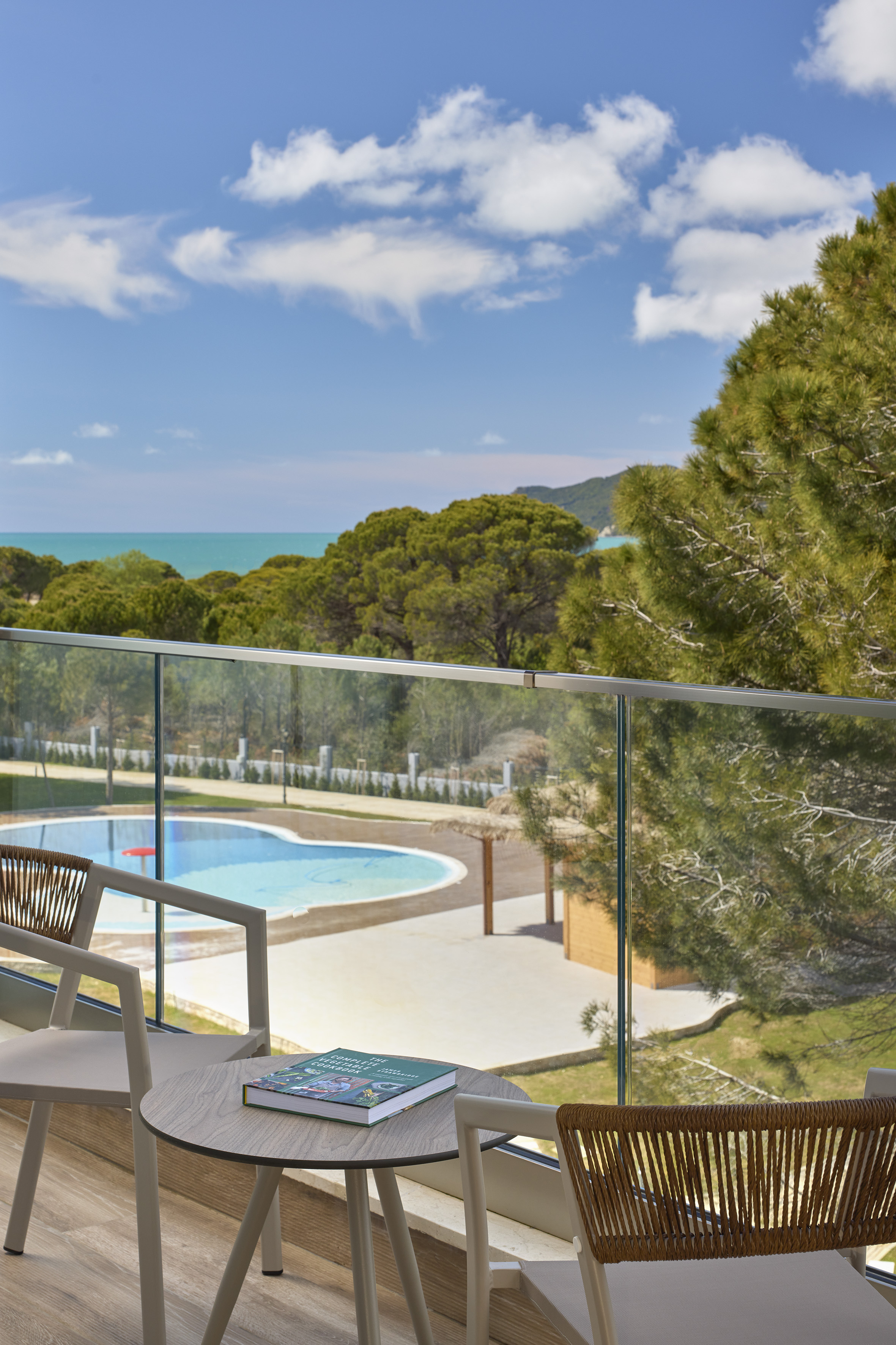 Balcony with chairs, table, book; overlooking pool, forest, and turquoise sea.