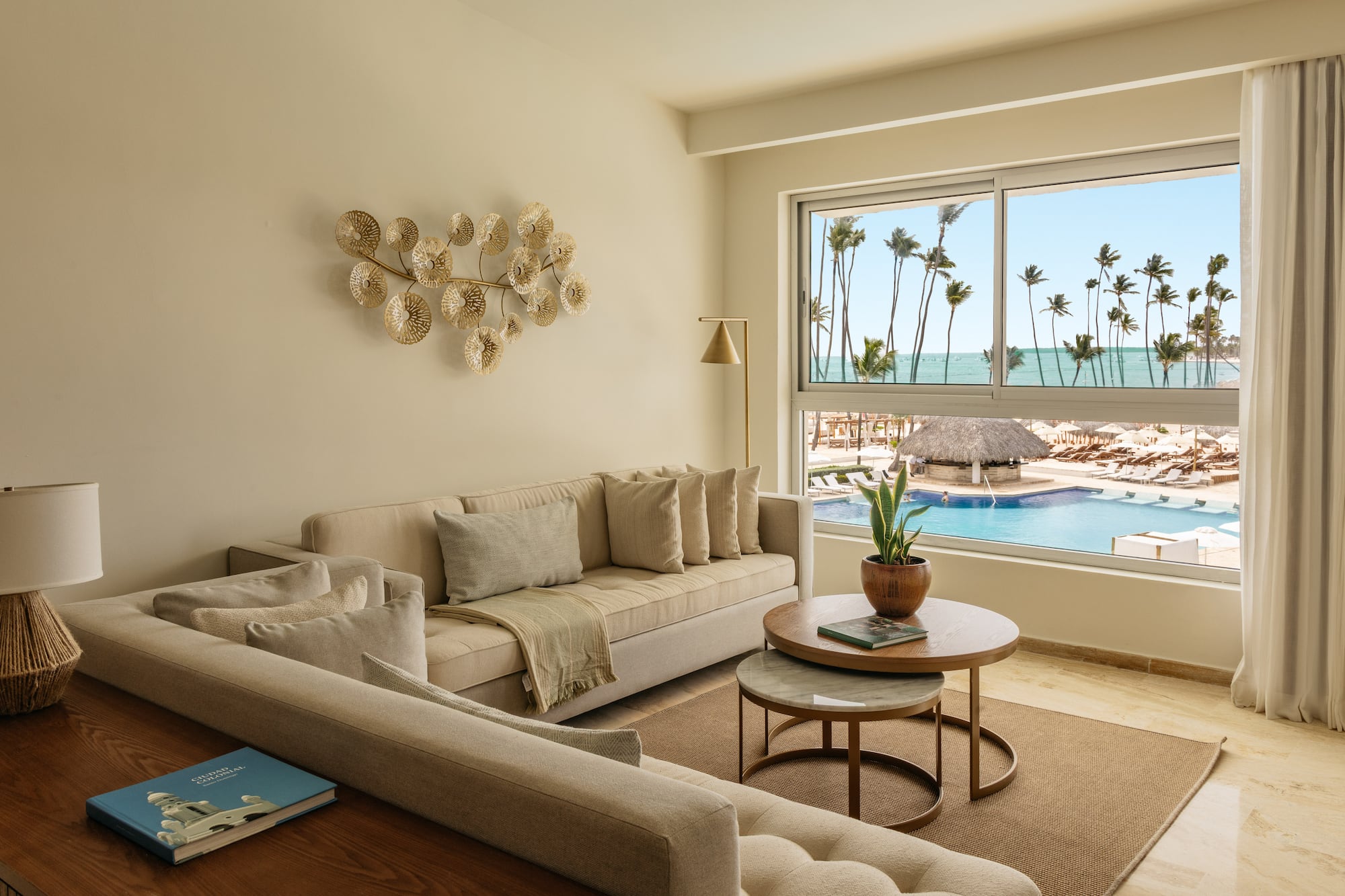Modern living room overlooking resort pool, ocean, and palm trees.