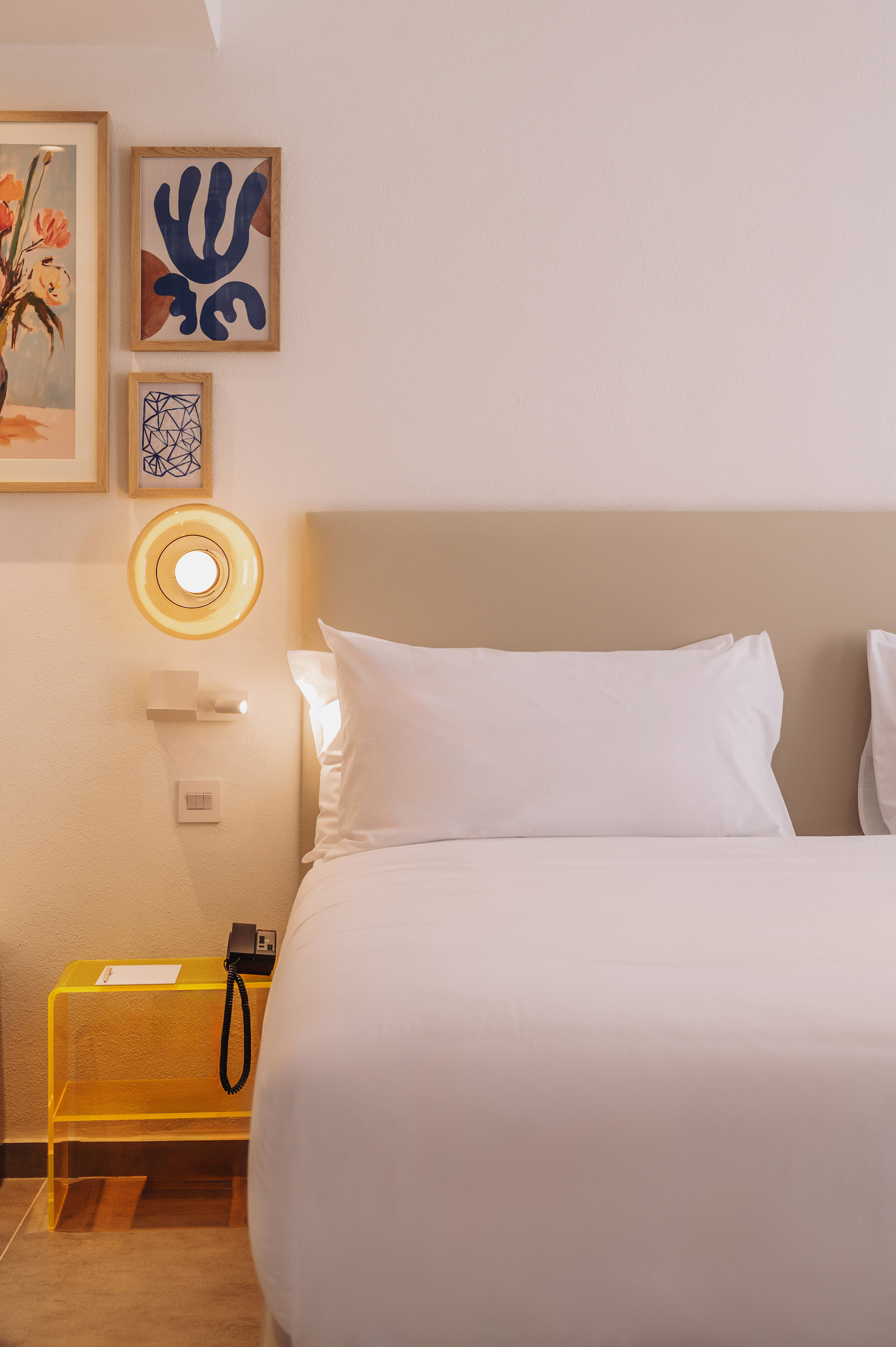 Modern hotel bedroom: white bed, yellow nightstand, abstract art, warm lamp.