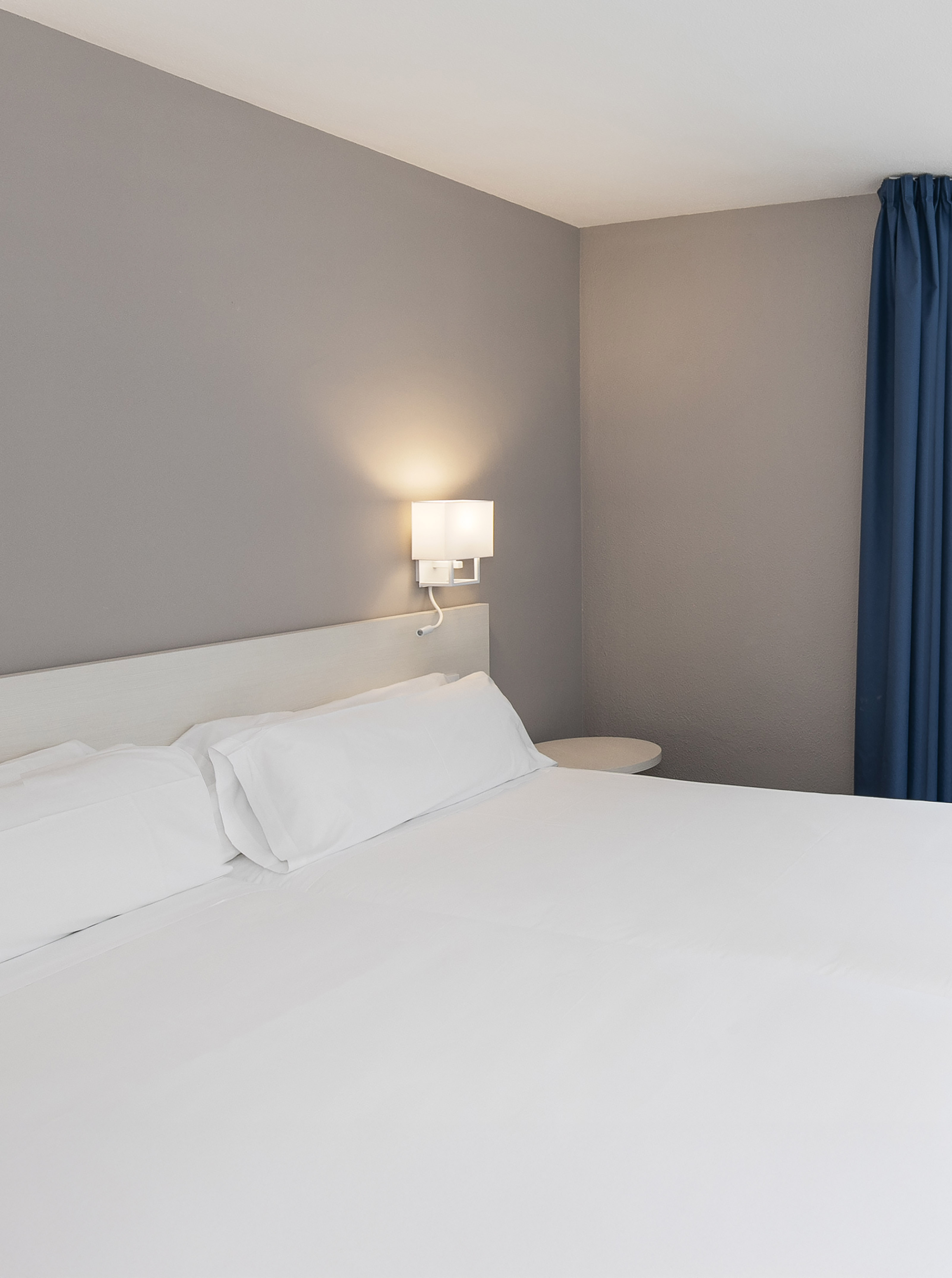 Modern hotel room with white bed, bedside lamp, grey walls, and blue curtain.