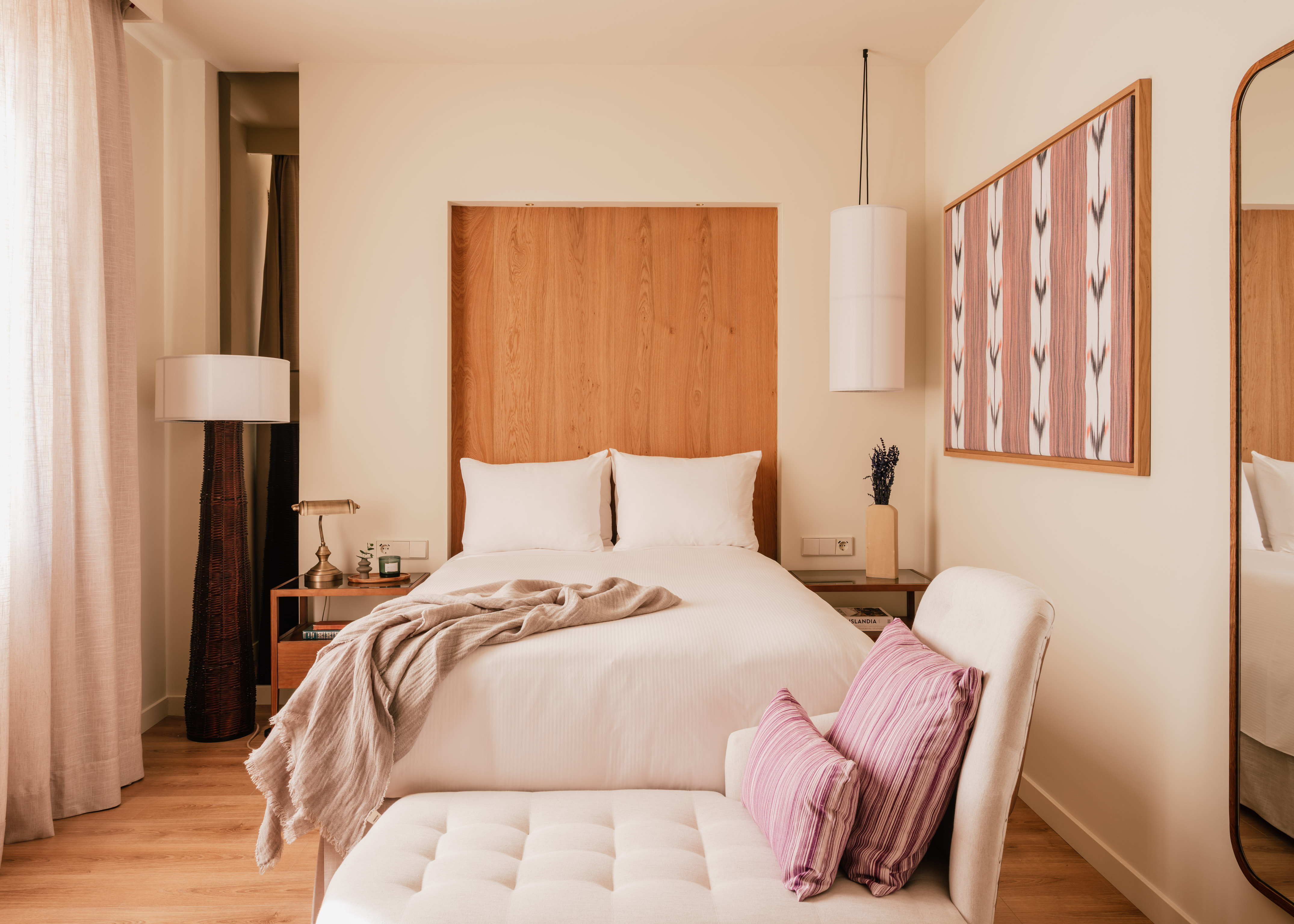Modern hotel room with white bed, wooden headboard, bench, and patterned art.