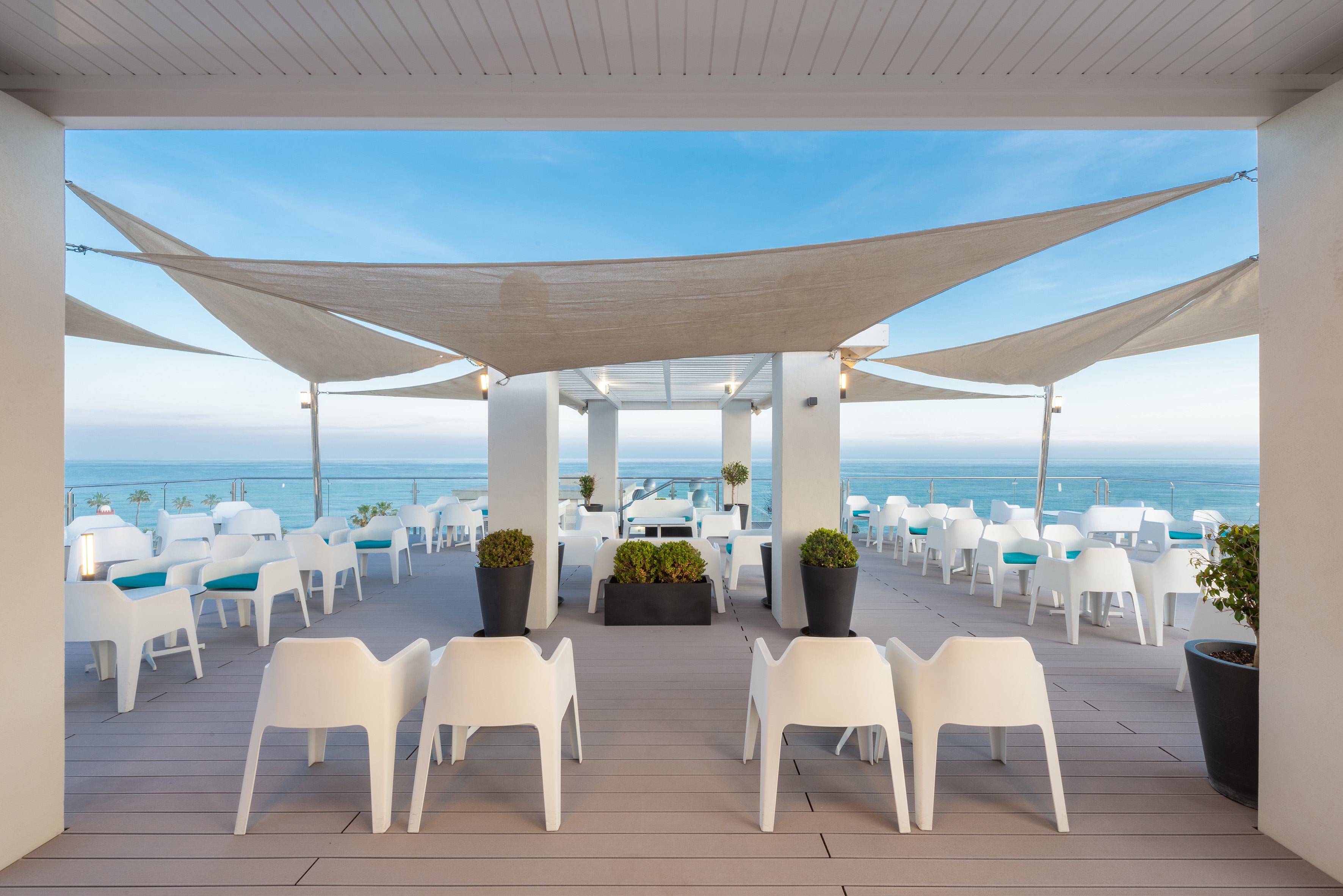 Outdoor restaurant patio with ocean view, white seating, and shade sails.