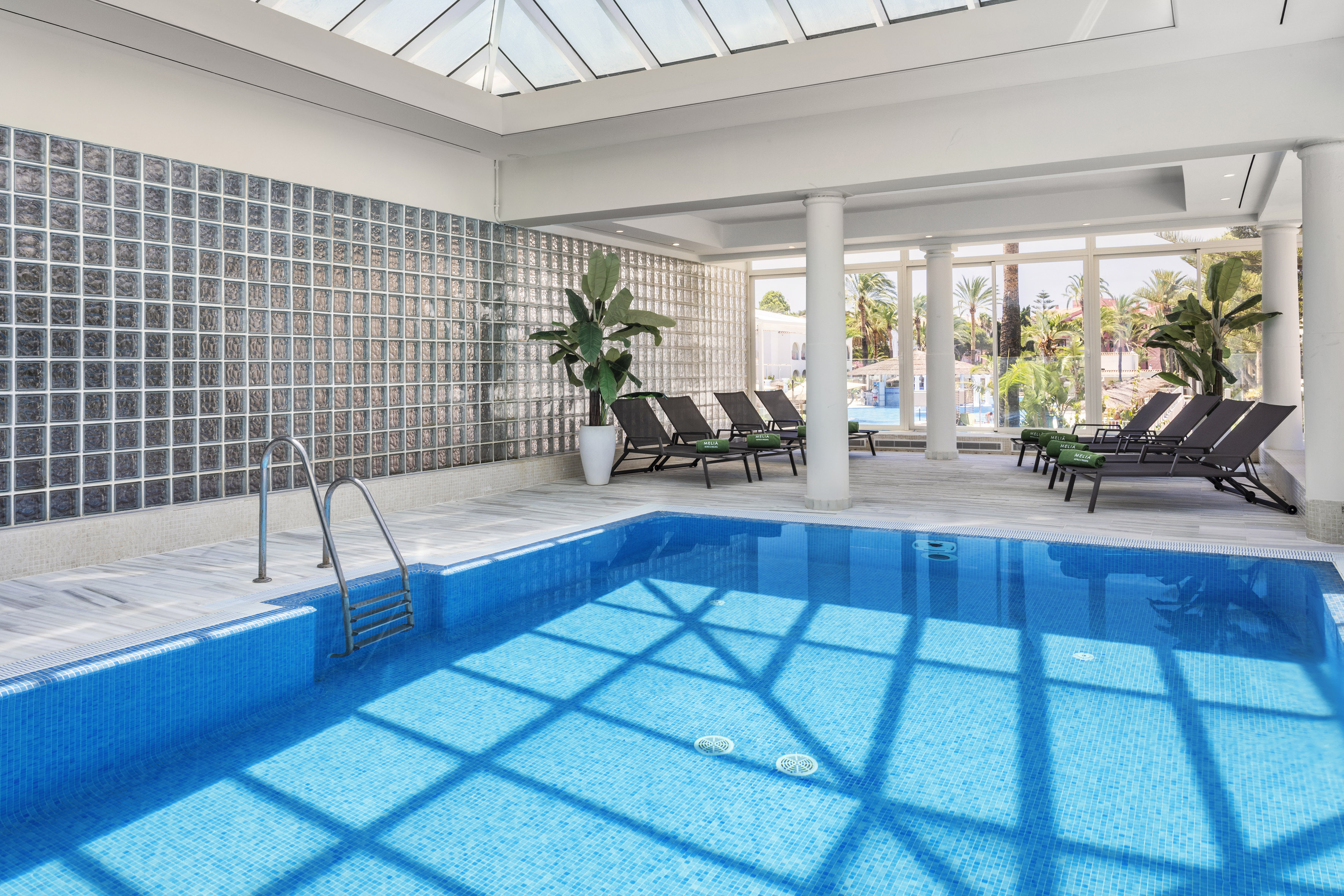 Bright indoor resort pool with blue tiles, glass block wall, and sun loungers.
