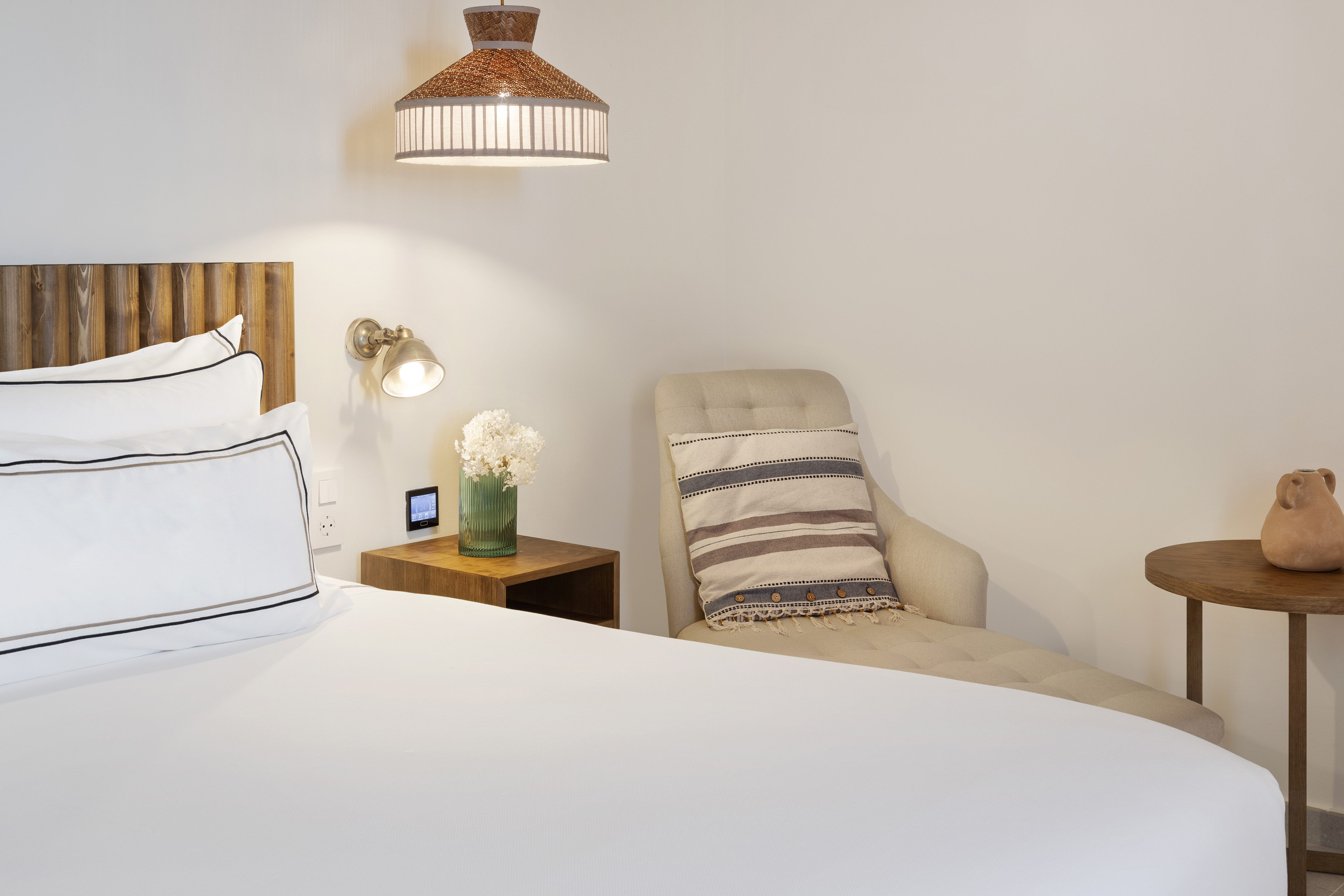 Modern hotel bedroom with white bed, wooden headboard, cozy chaise, and rattan lamp.