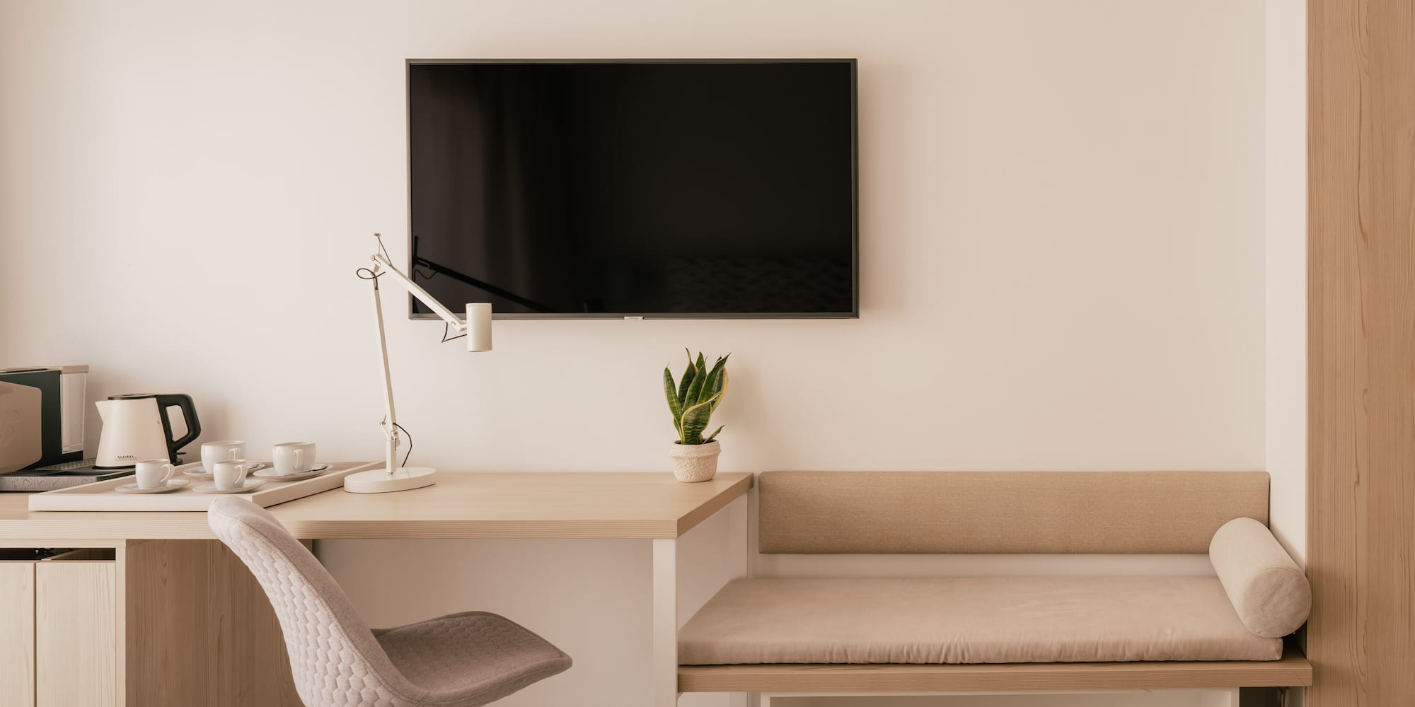 Modern hotel room with desk, wall TV, coffee station, and bench seating.