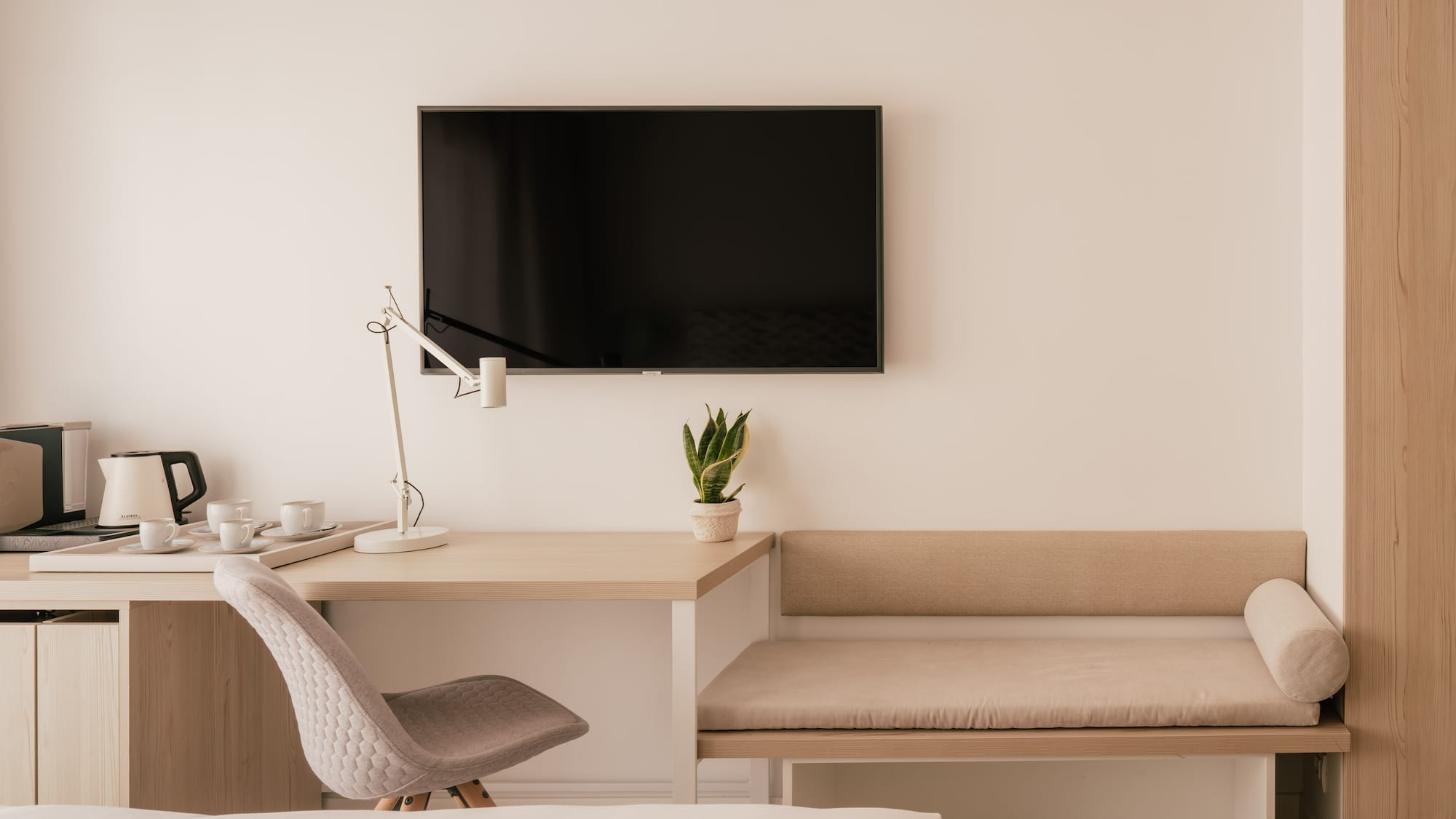 Modern hotel room with desk, wall TV, coffee station, and bench seating.