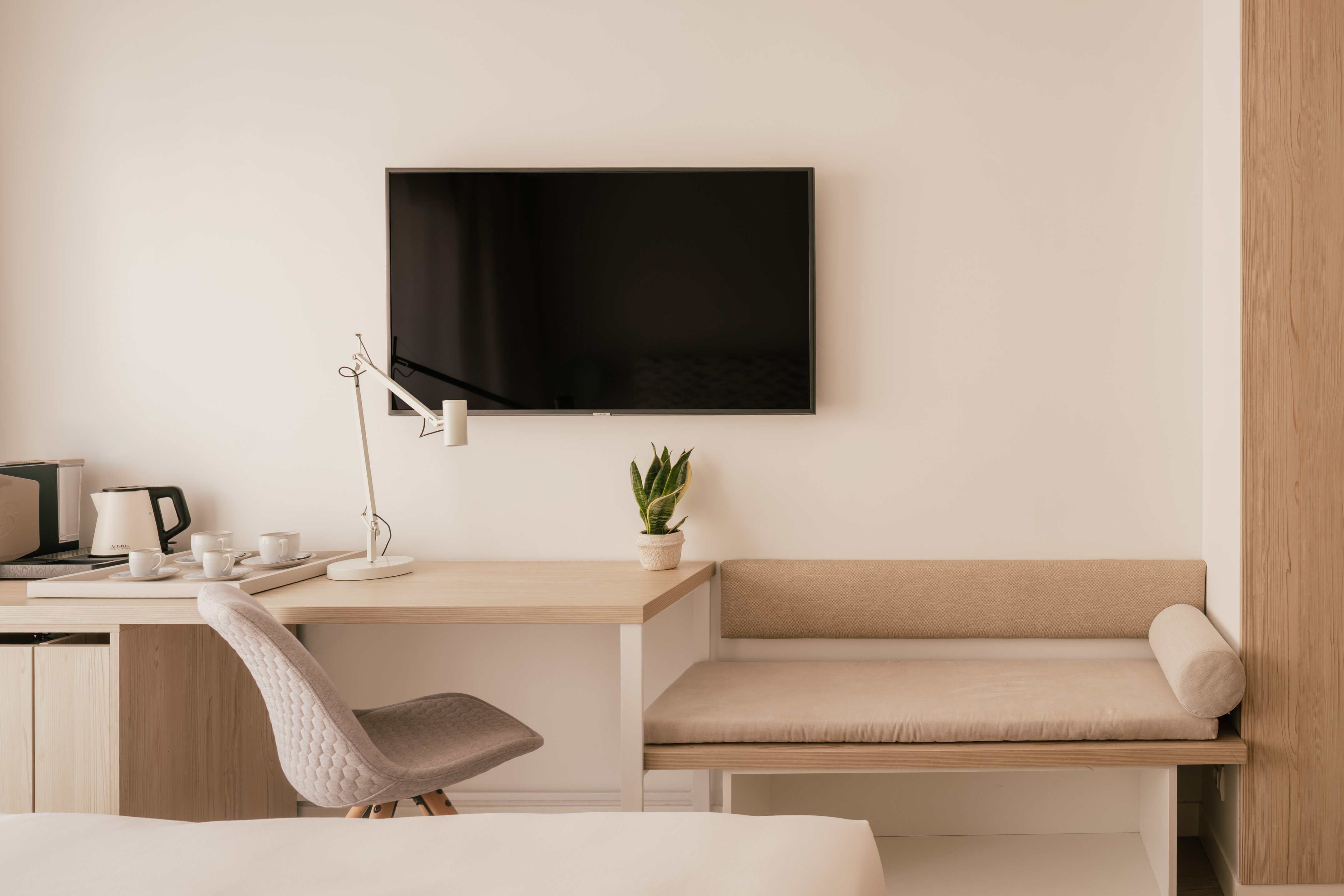 Modern hotel room with desk, wall TV, coffee station, and bench seating.