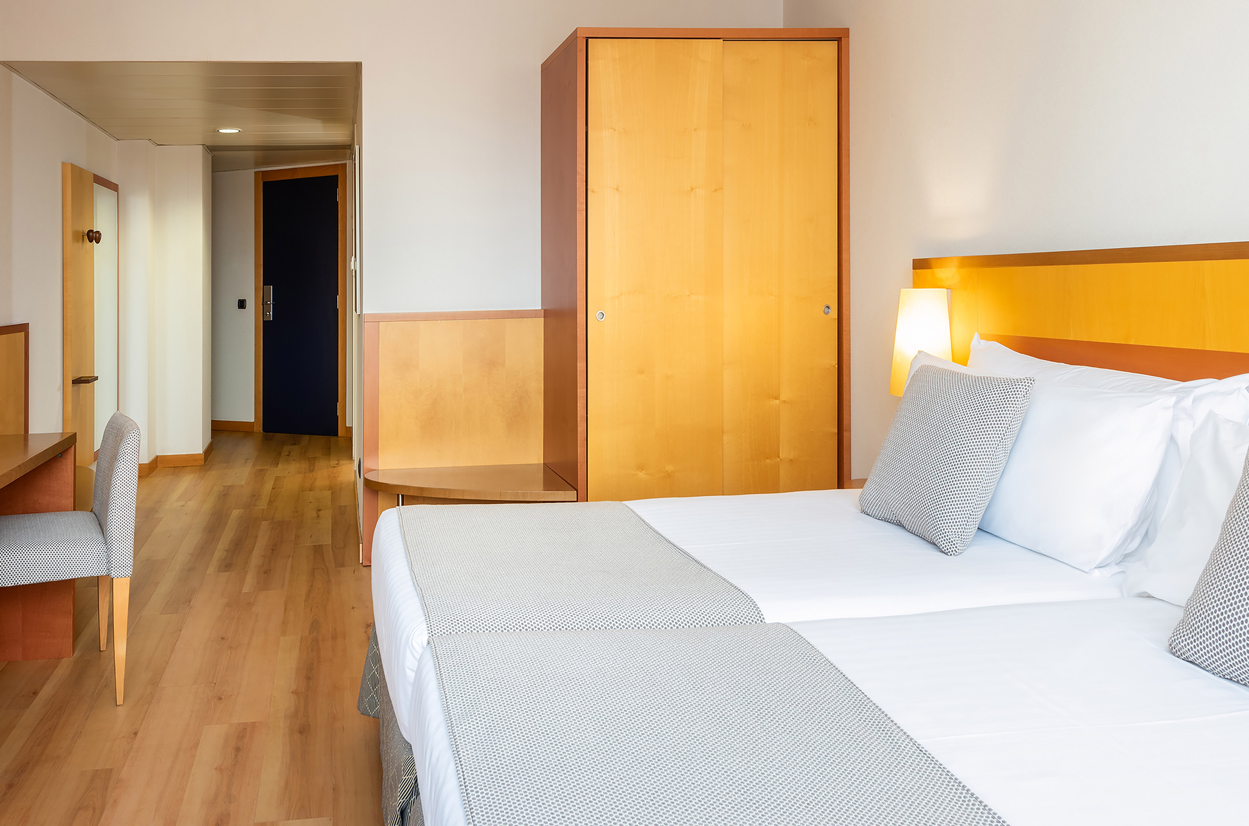 Hotel room with two beds, wooden wardrobe, desk, and light-colored wooden floor.