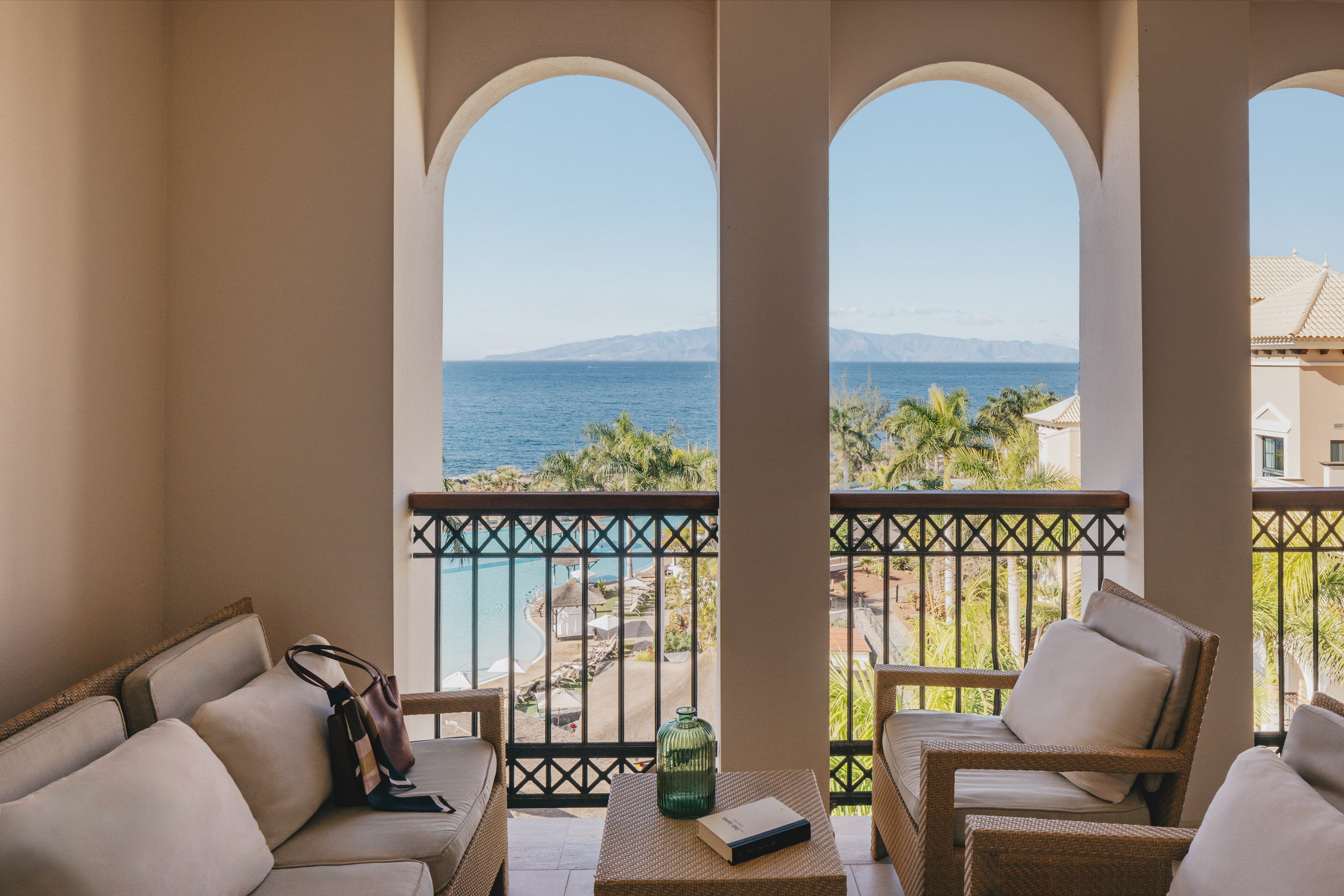Oceanfront balcony with arched sea view, mountains, palm trees, and pool.
