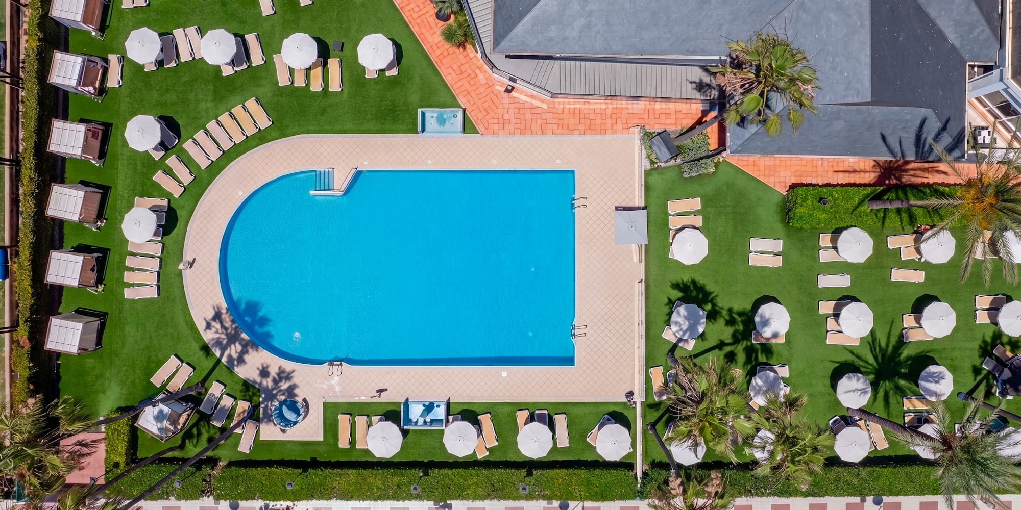 Aerial view hotel pool, sun loungers, umbrellas, green lawn, palm trees.