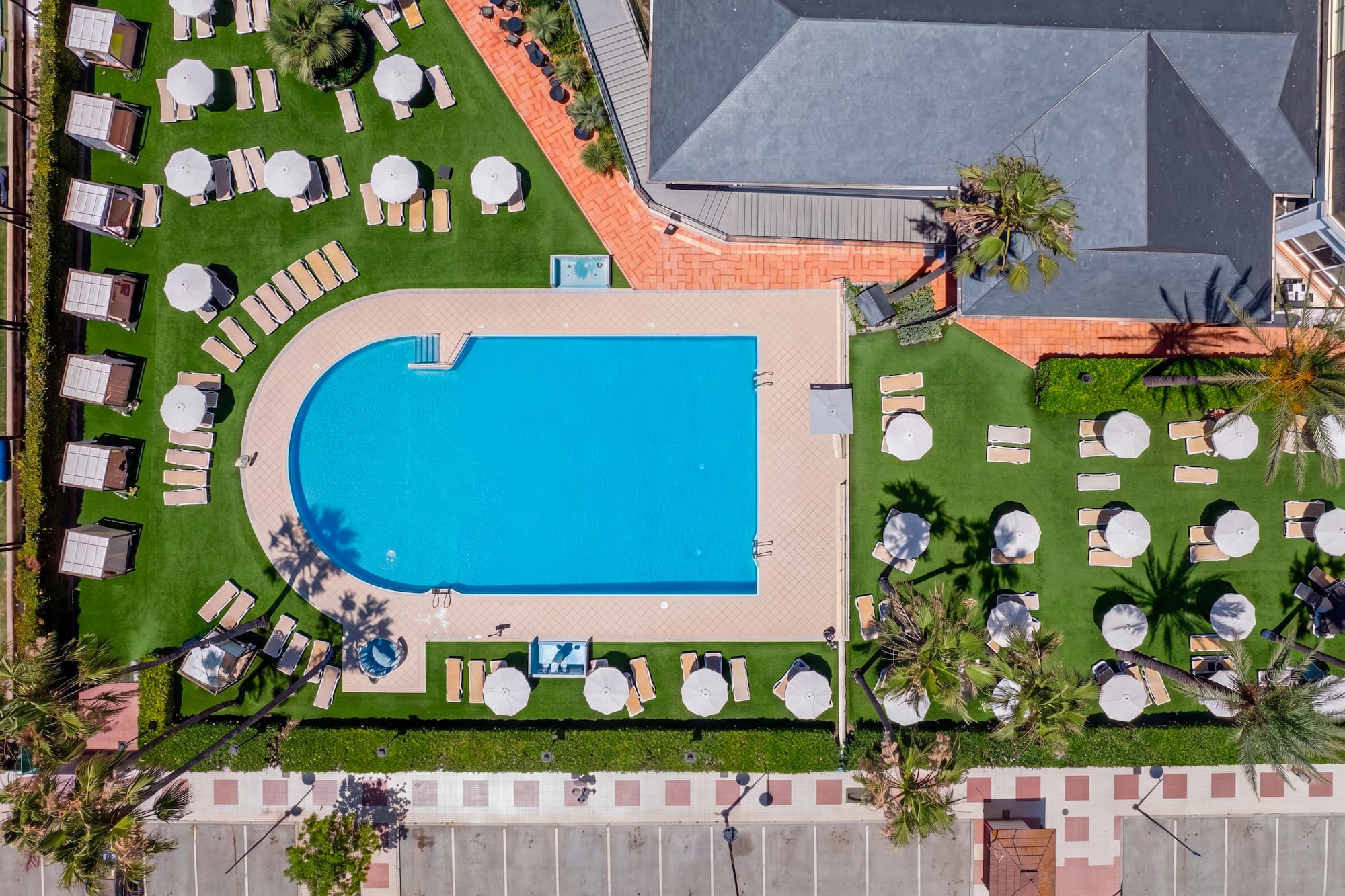 Aerial view hotel pool, sun loungers, umbrellas, green lawn, palm trees.