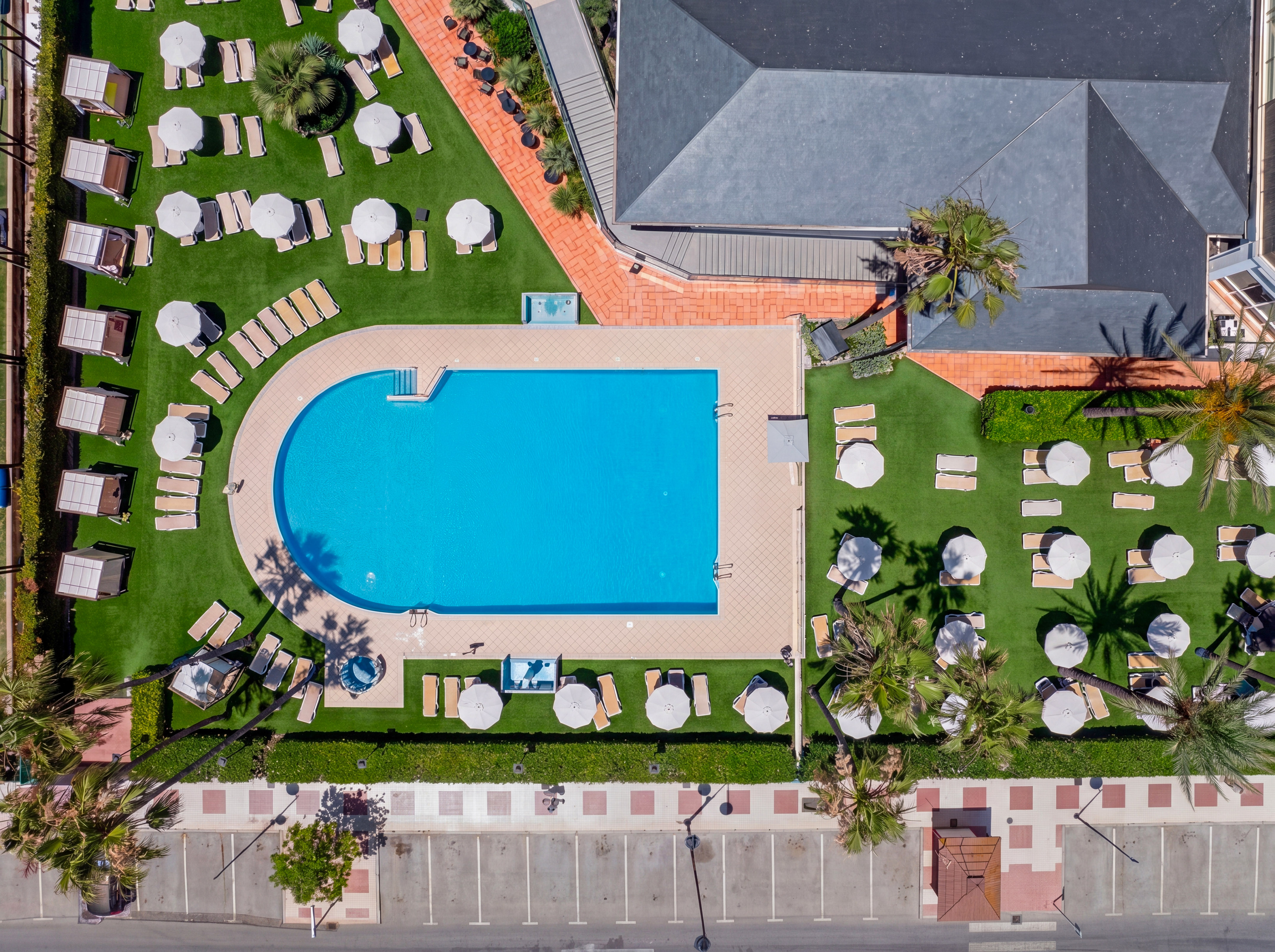 Aerial view hotel pool, sun loungers, umbrellas, green lawn, palm trees.