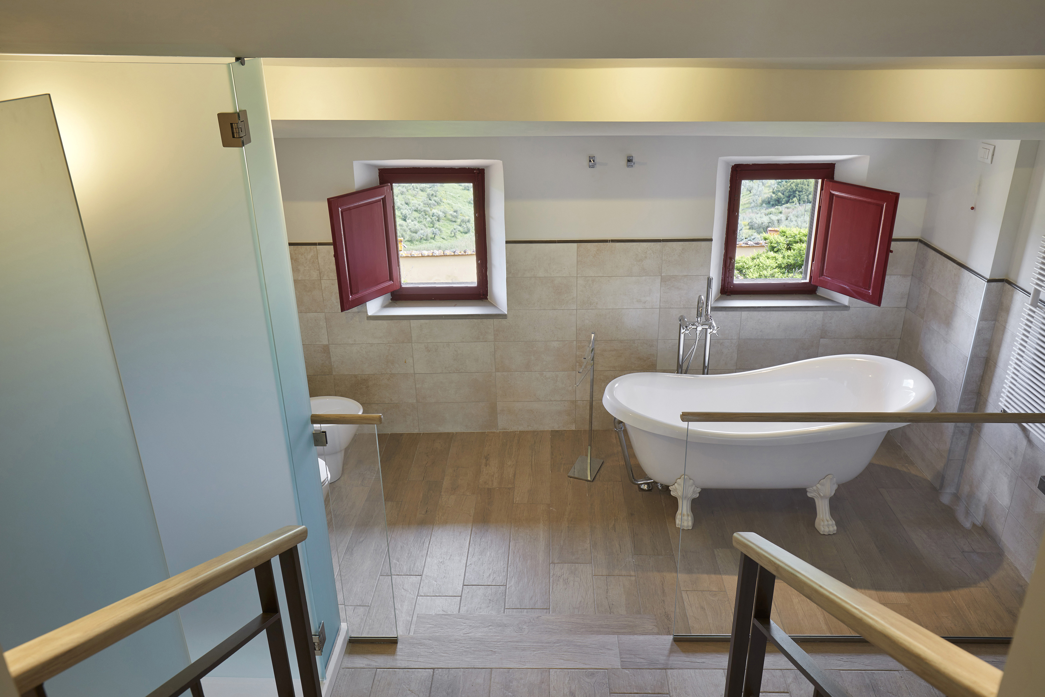 Rustic bathroom with clawfoot tub, red-shuttered windows, and hillside view.