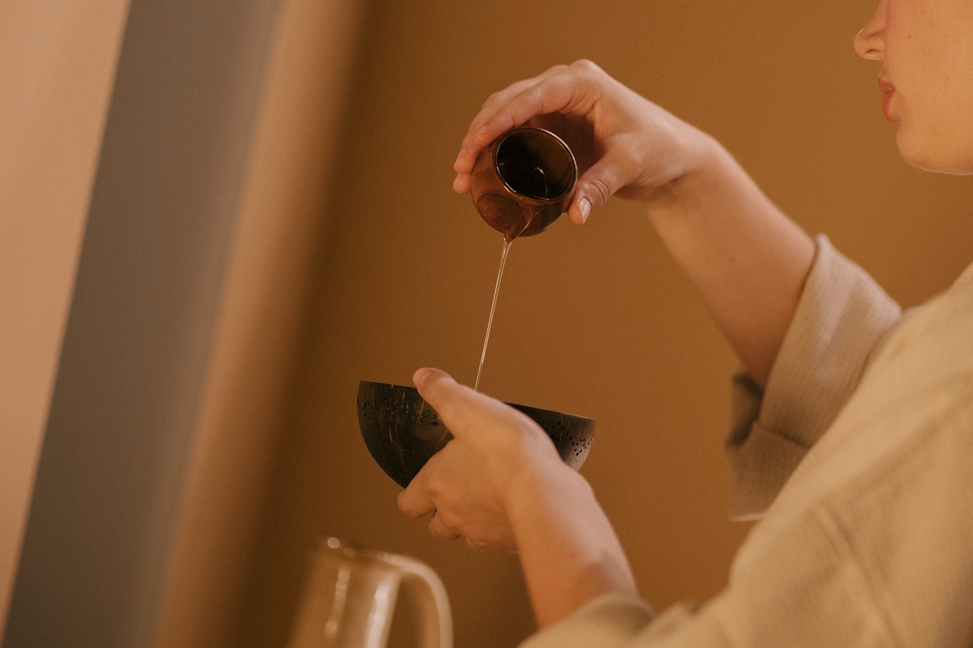 Woman's hands pouring oil from a small cup into a natural coconut bowl for a spa ritual.