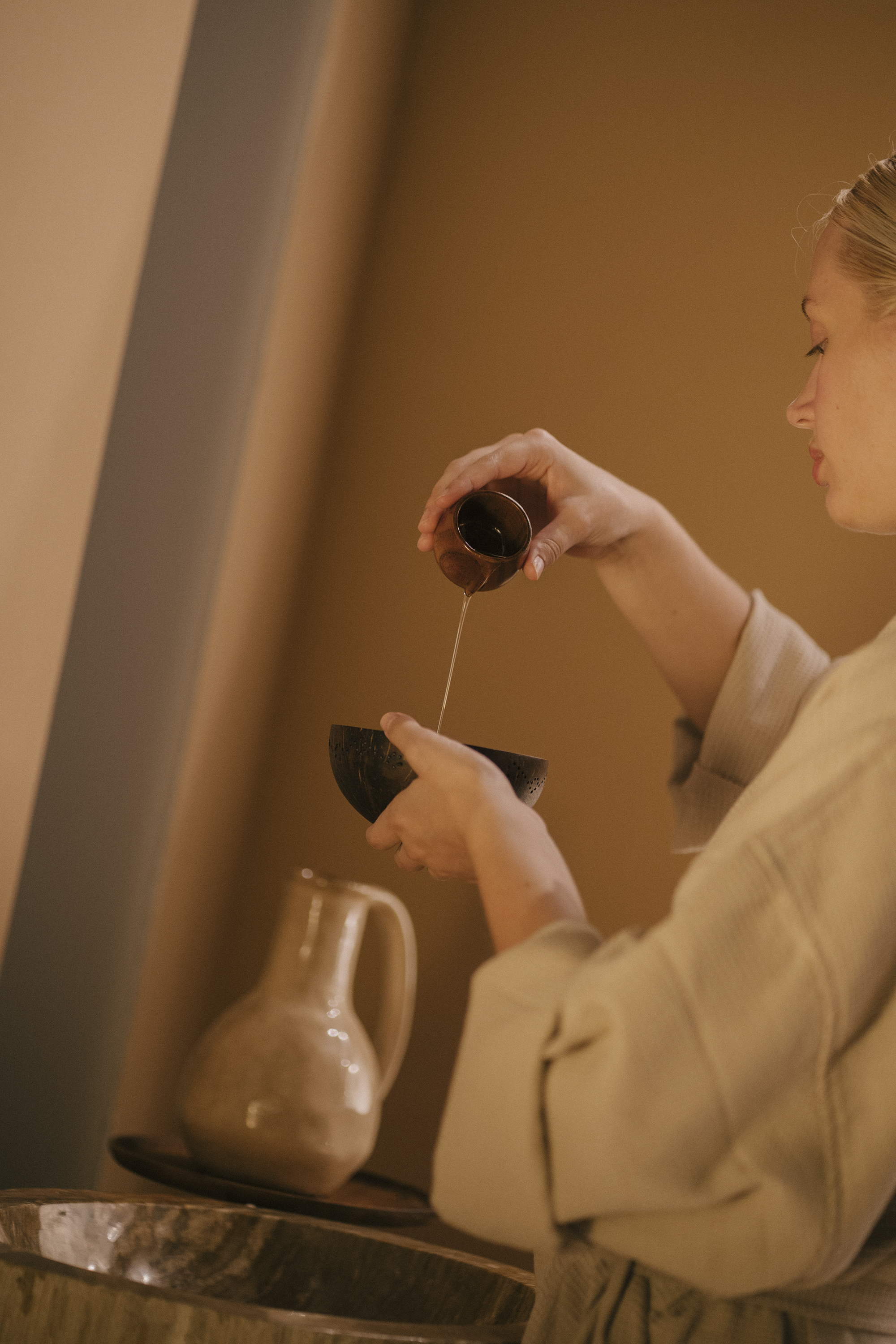 Woman's hands pouring oil from a small cup into a natural coconut bowl for a spa ritual.
