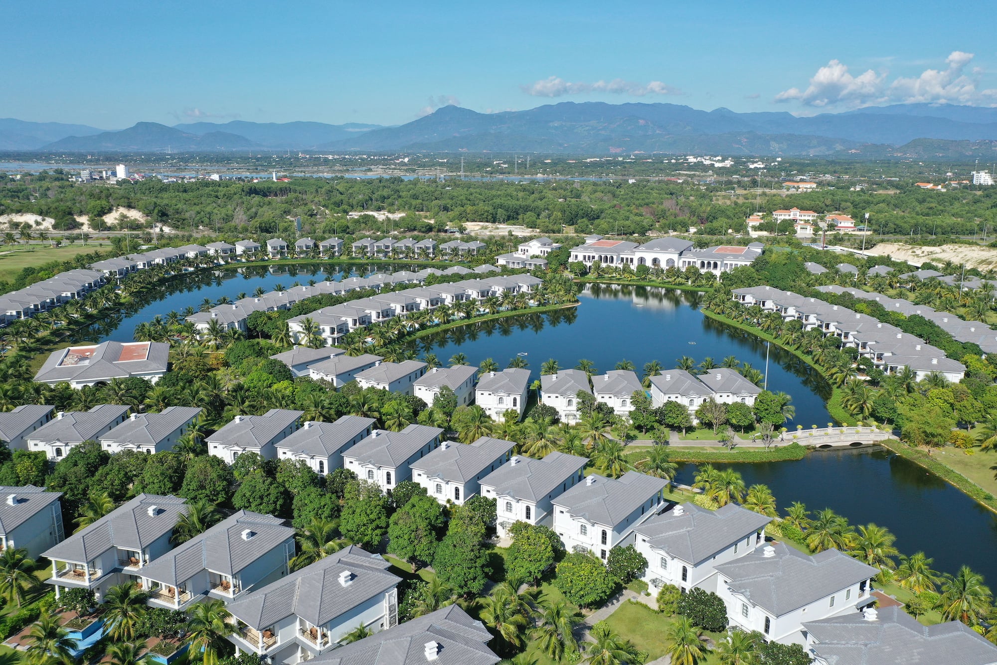 Aerial view: modern waterfront villas lining a lake, tropical trees, mountains.