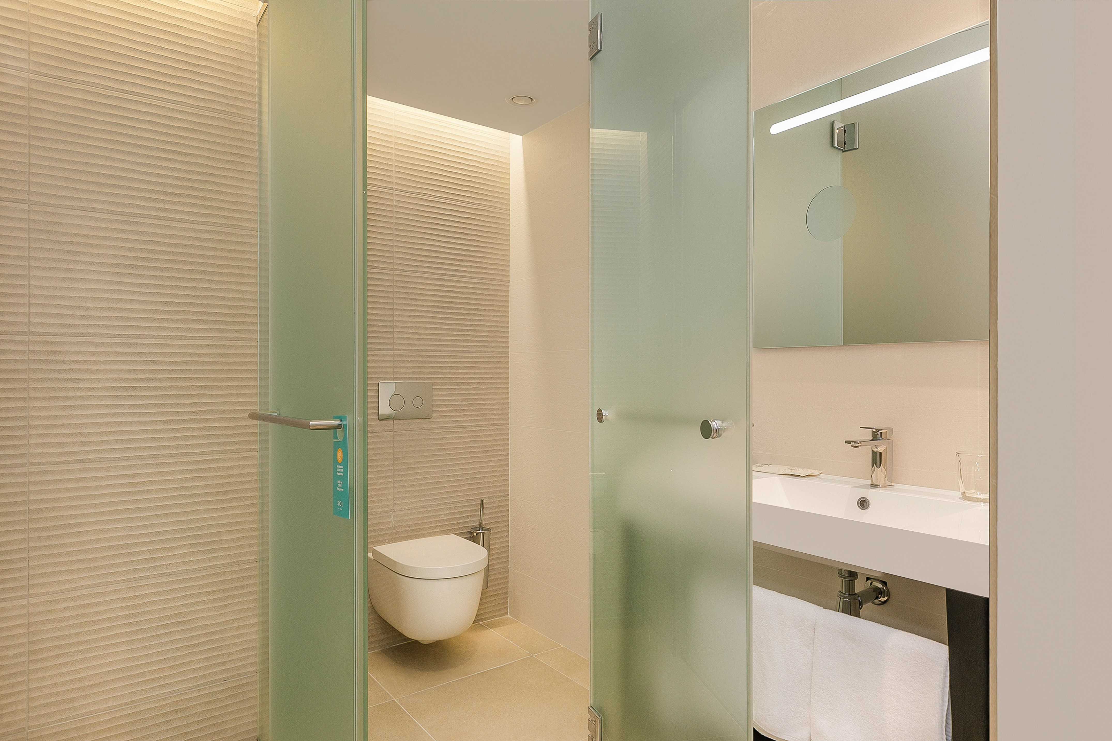 Modern hotel bathroom with wall-mounted toilet, sink, and frosted glass shower.