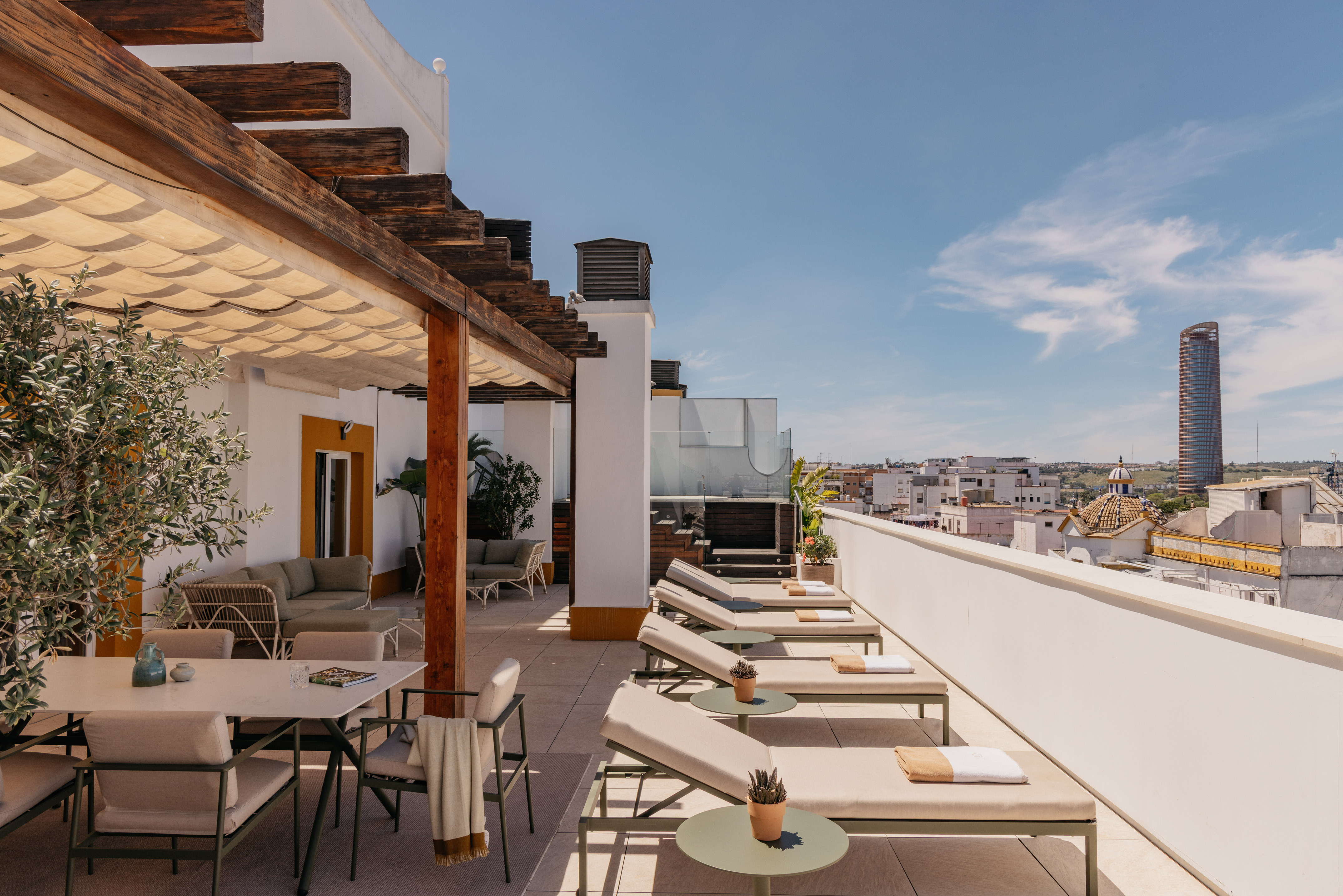Terraza de azotea soleada con pérgola, tumbonas, mesa y vistas a Sevilla.