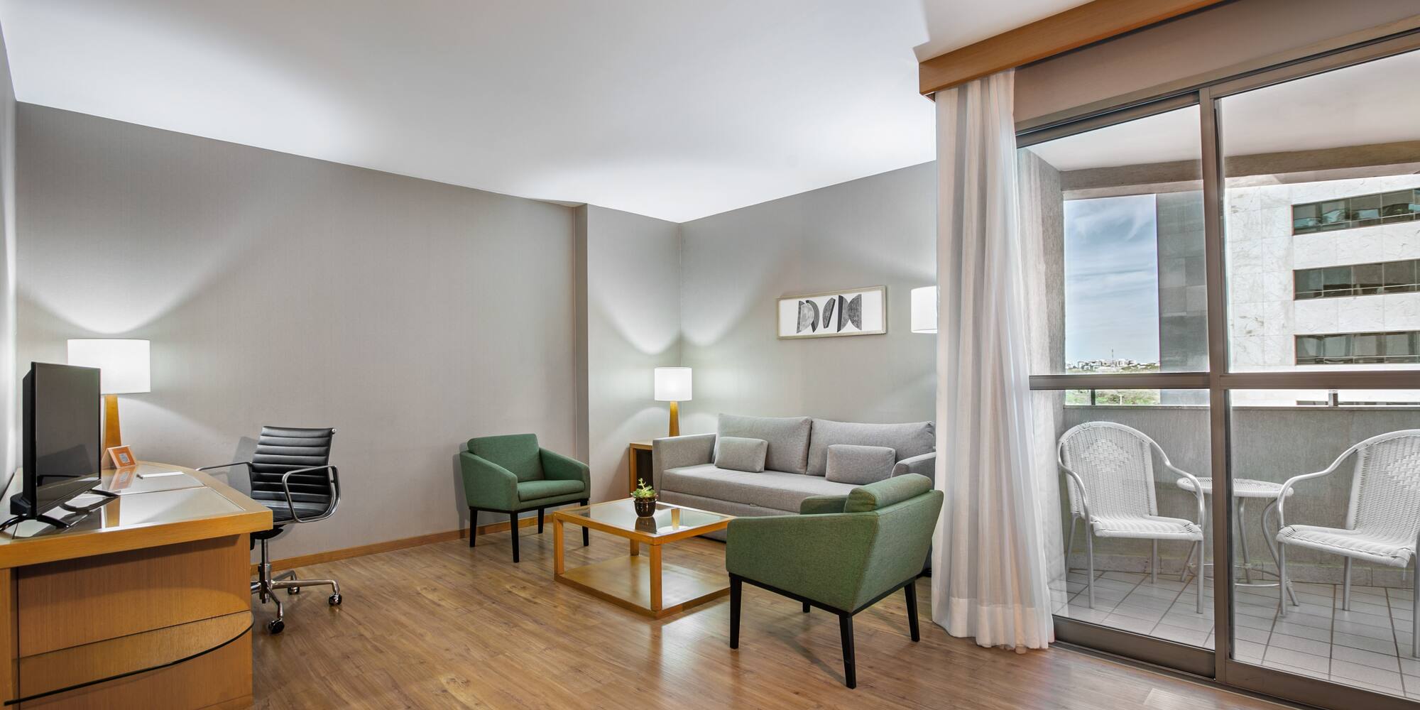 Modern hotel suite living room with gray sofa, green chairs, desk, and balcony.