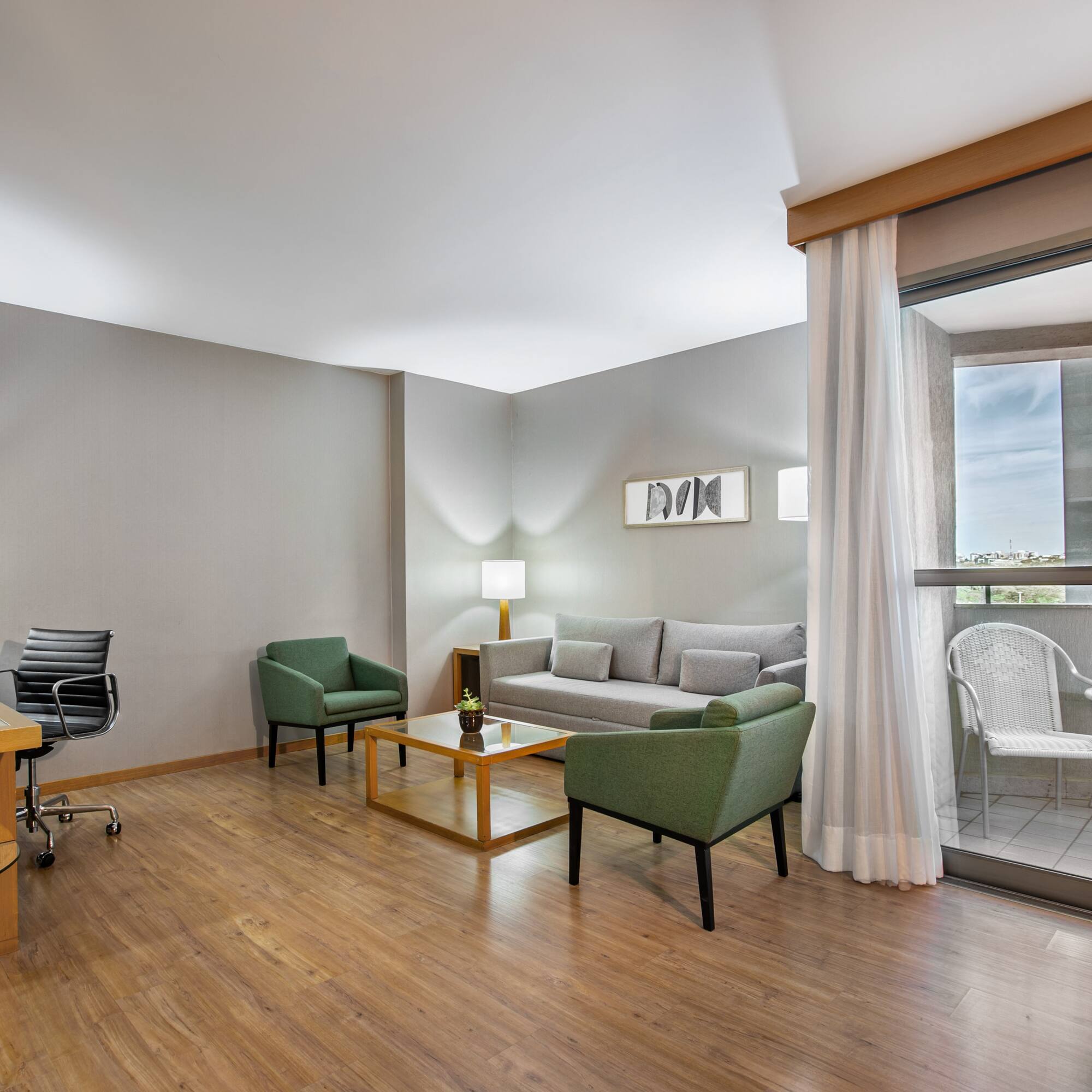 Modern hotel suite living room with gray sofa, green chairs, desk, and balcony.