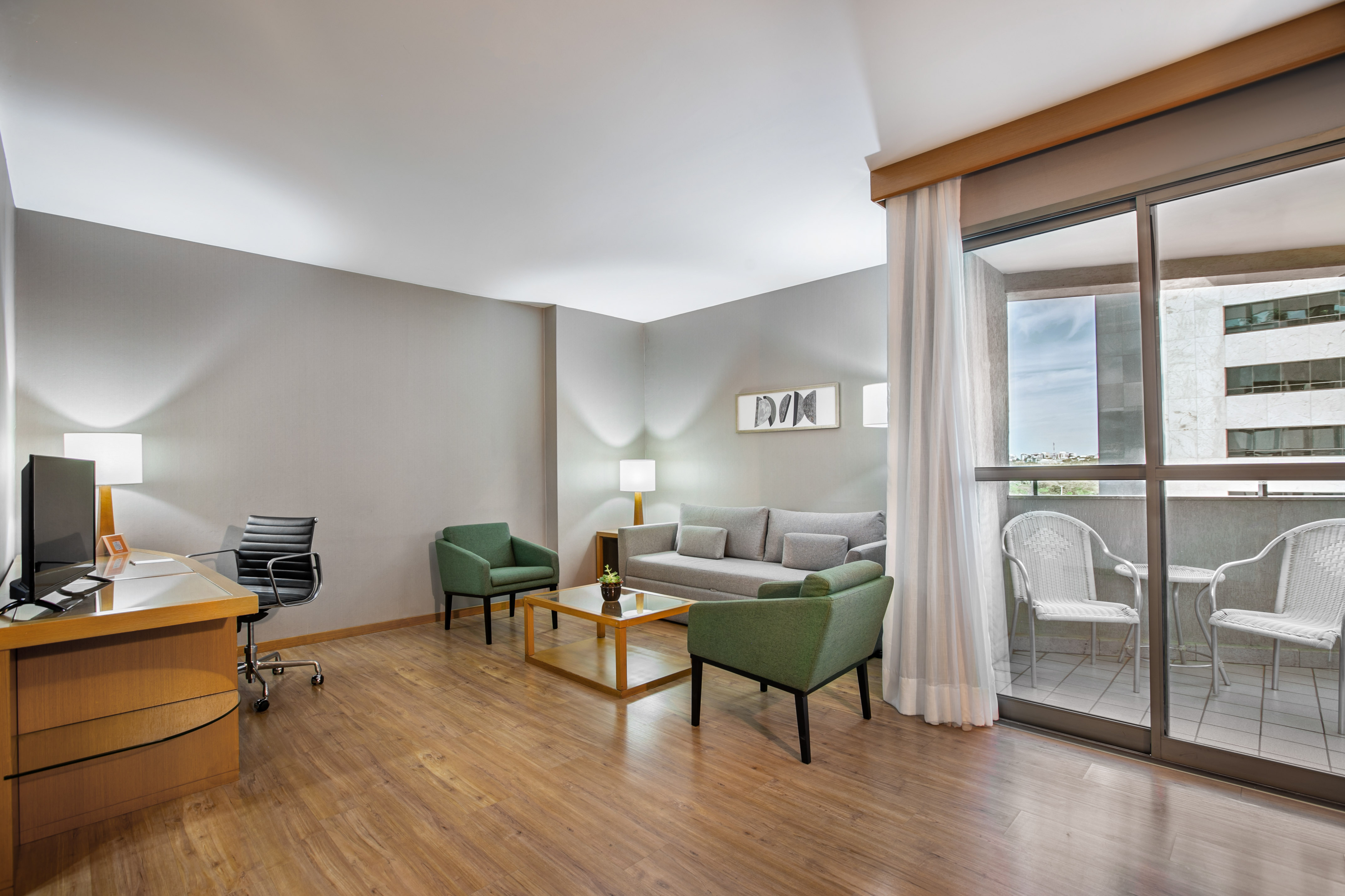 Modern hotel suite living room with gray sofa, green chairs, desk, and balcony.