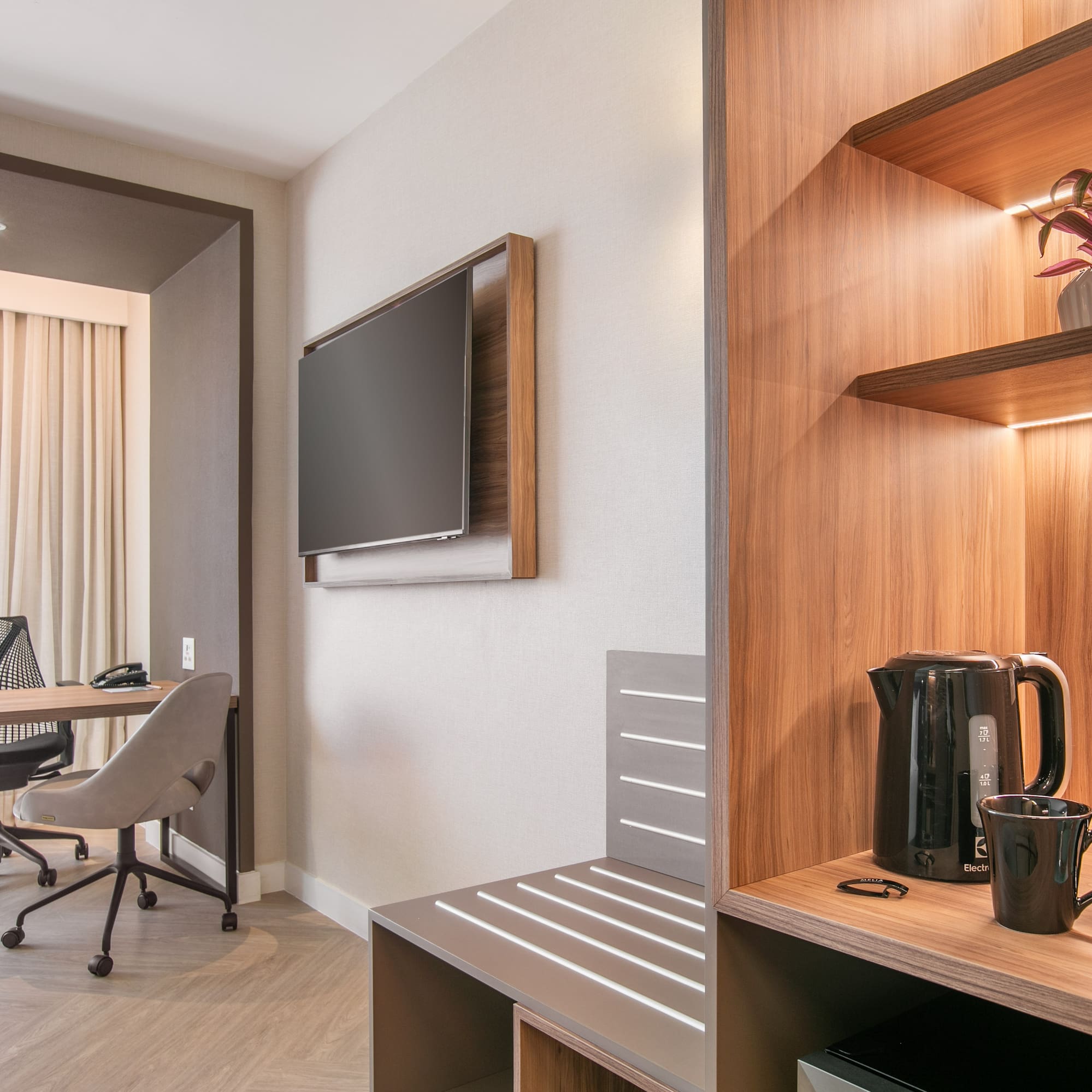 Modern hotel room with workspace, TV, and illuminated coffee/tea station.
