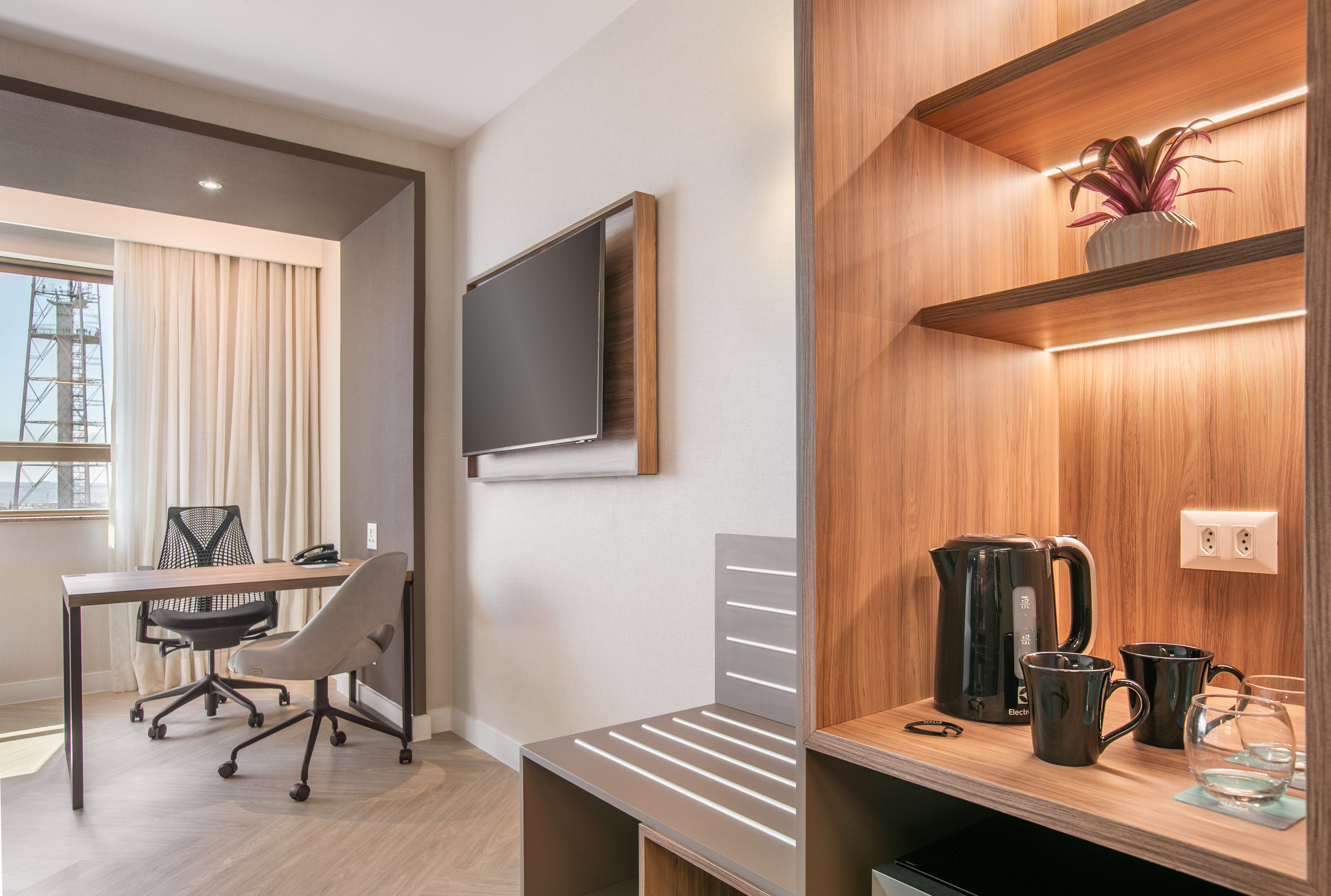 Modern hotel room with workspace, wall TV, and beverage station.