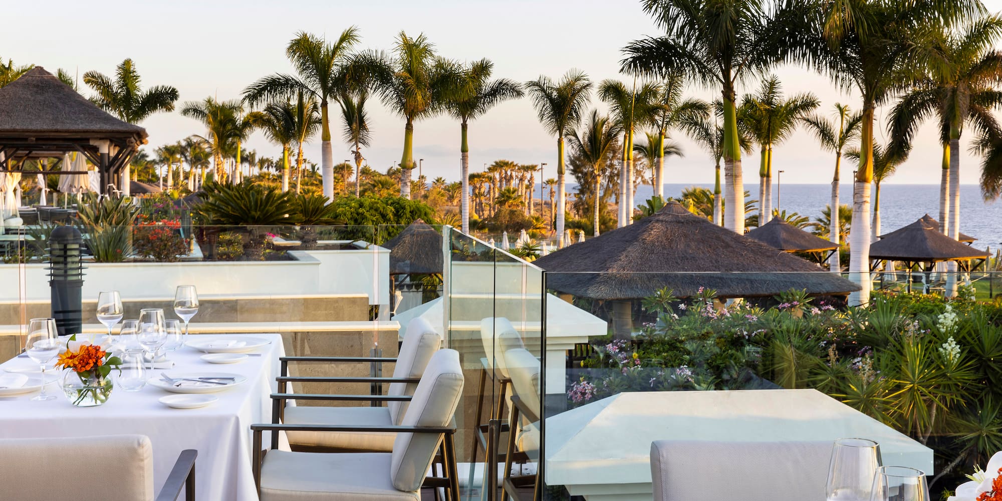 a table set up with white plates and chairs on a balcony overlooking a body of water