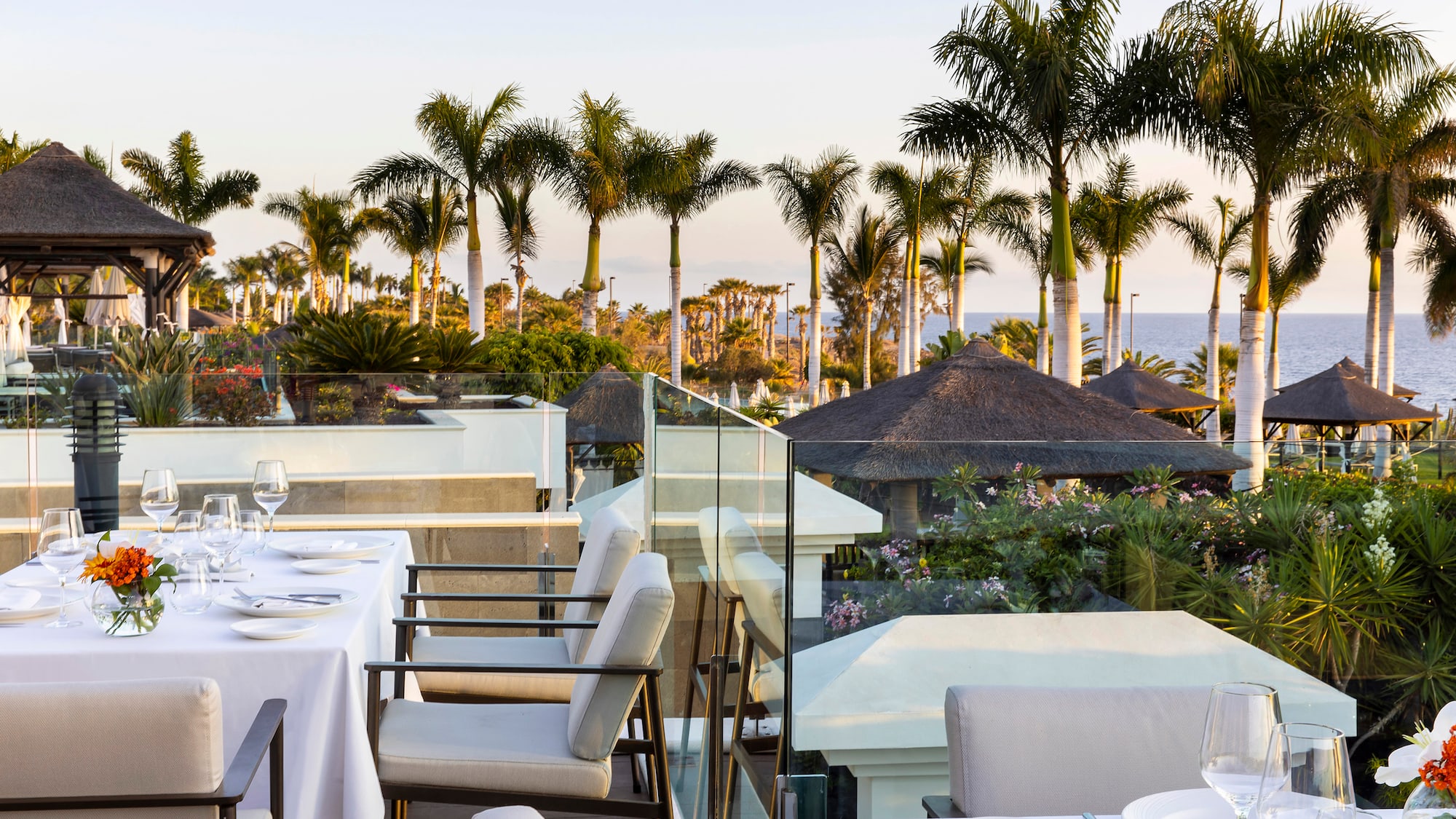 a table set up with white plates and chairs on a balcony overlooking a body of water