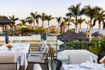 a table set up with white plates and chairs on a balcony overlooking a body of water