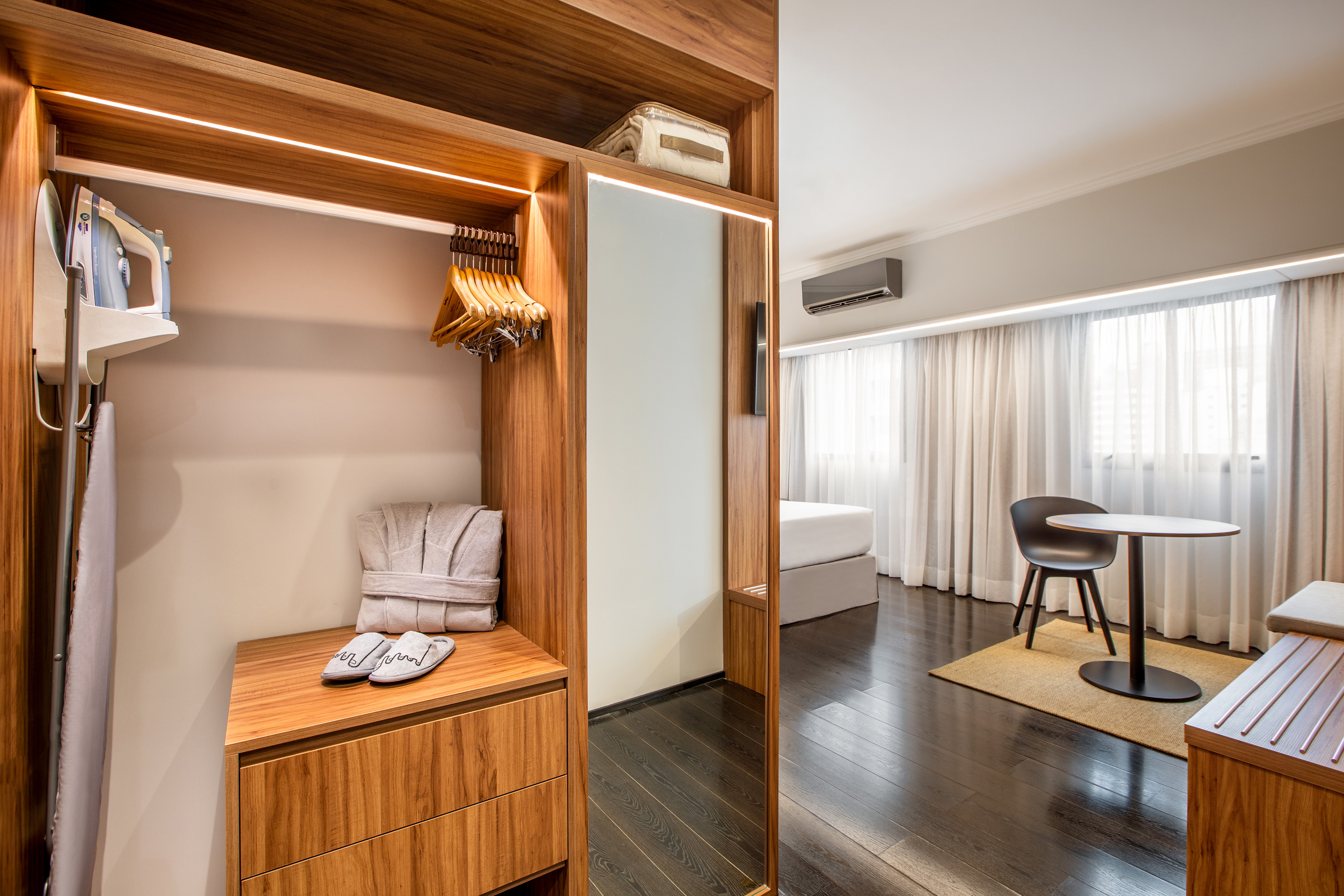 Modern hotel room with open wooden closet, iron, bathrobe, mirror, and bed.