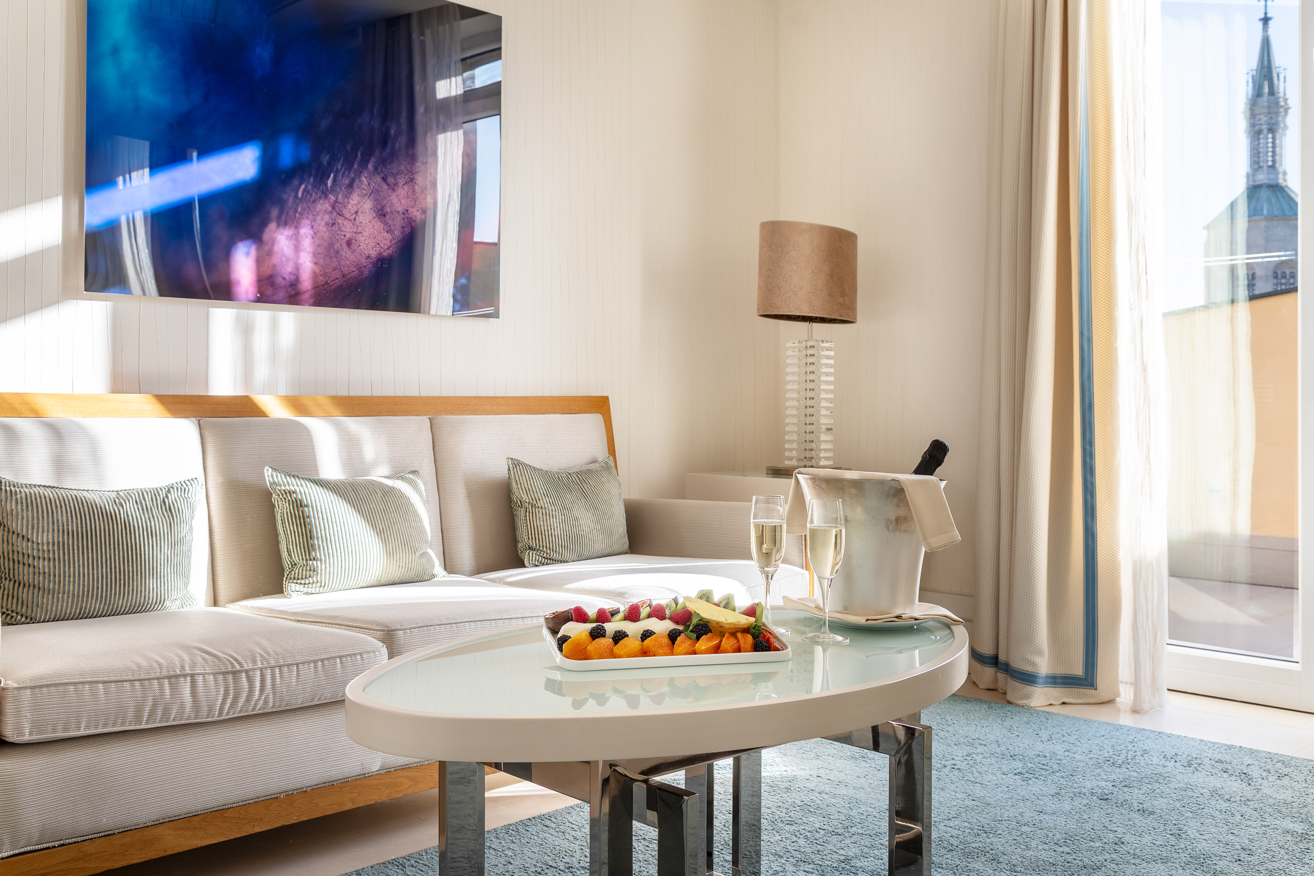 Hotel suite lounge with fruit platter, champagne, modern sofa, and city view.