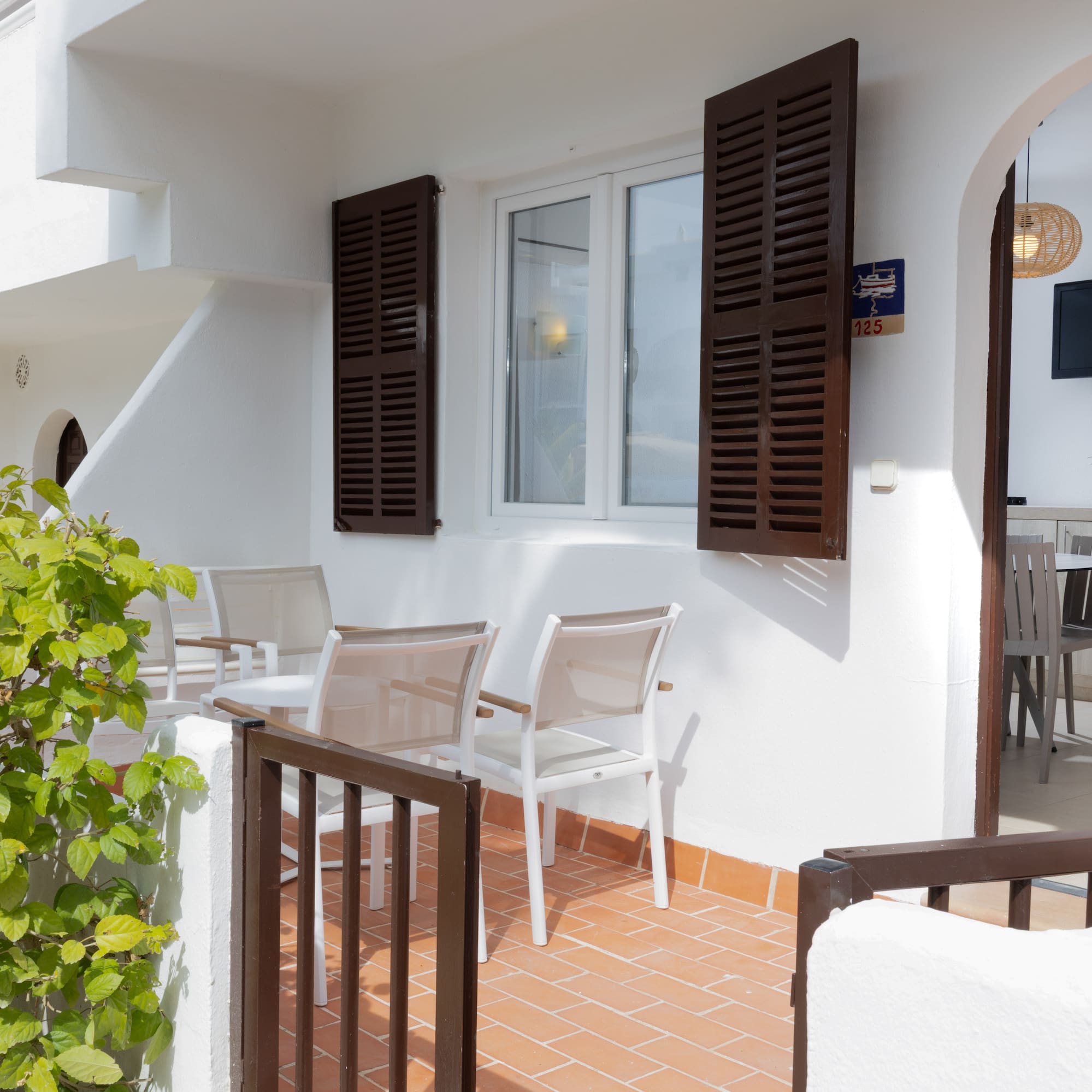 Bright apartment patio with outdoor seating, green bush, and room interior view.
