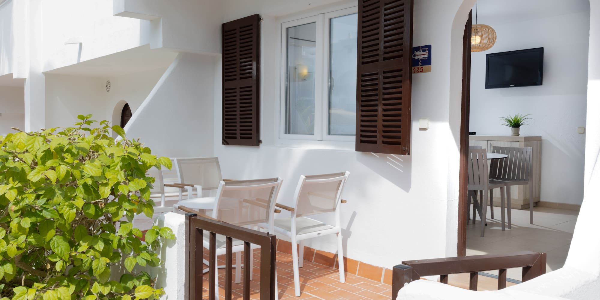 Bright apartment patio with outdoor seating, green bush, and room interior view.